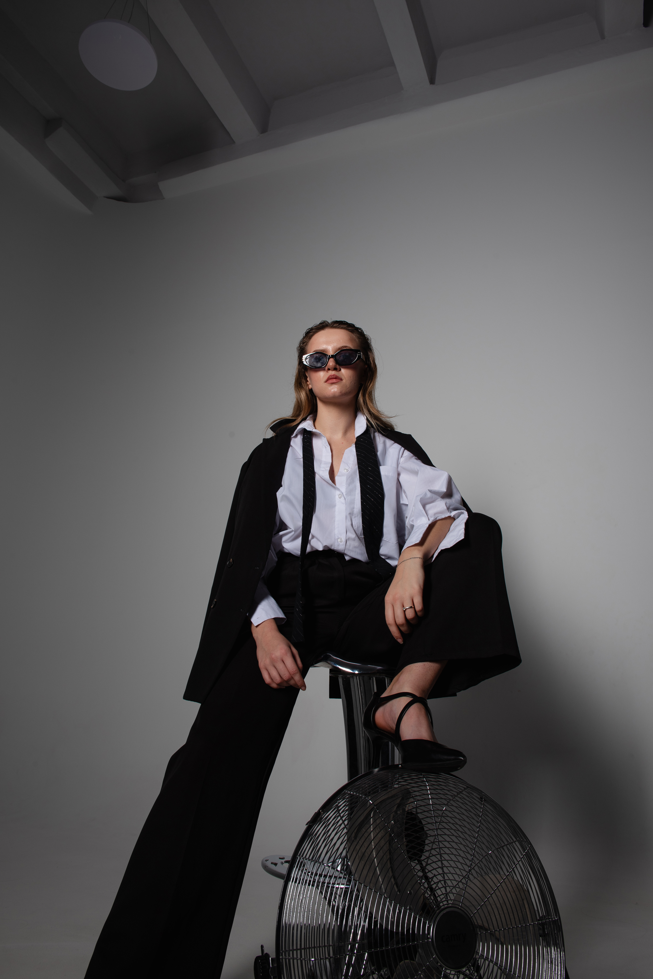 Fashion portrait before final retouching, showing a woman in tailored black and white clothing seated in a minimal studio with a large floor fan