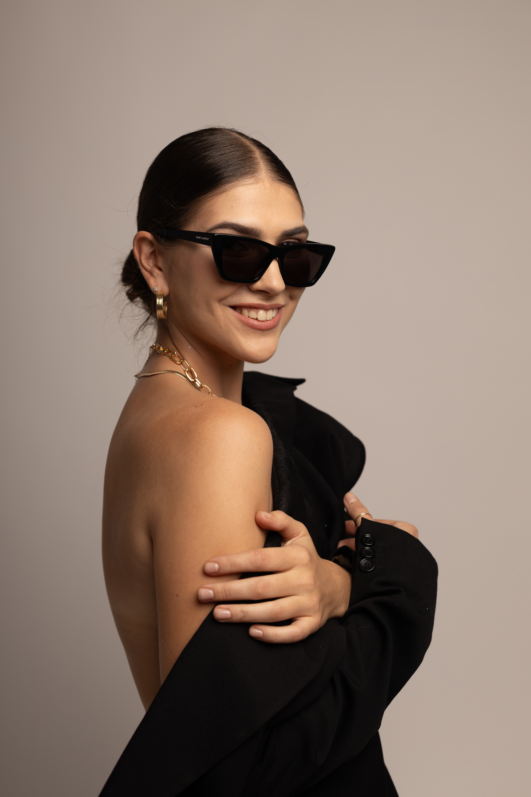 Fashion beauty portrait before final retouching, showing a woman in black sunglasses and a dark off-shoulder outfit against a warm neutral background