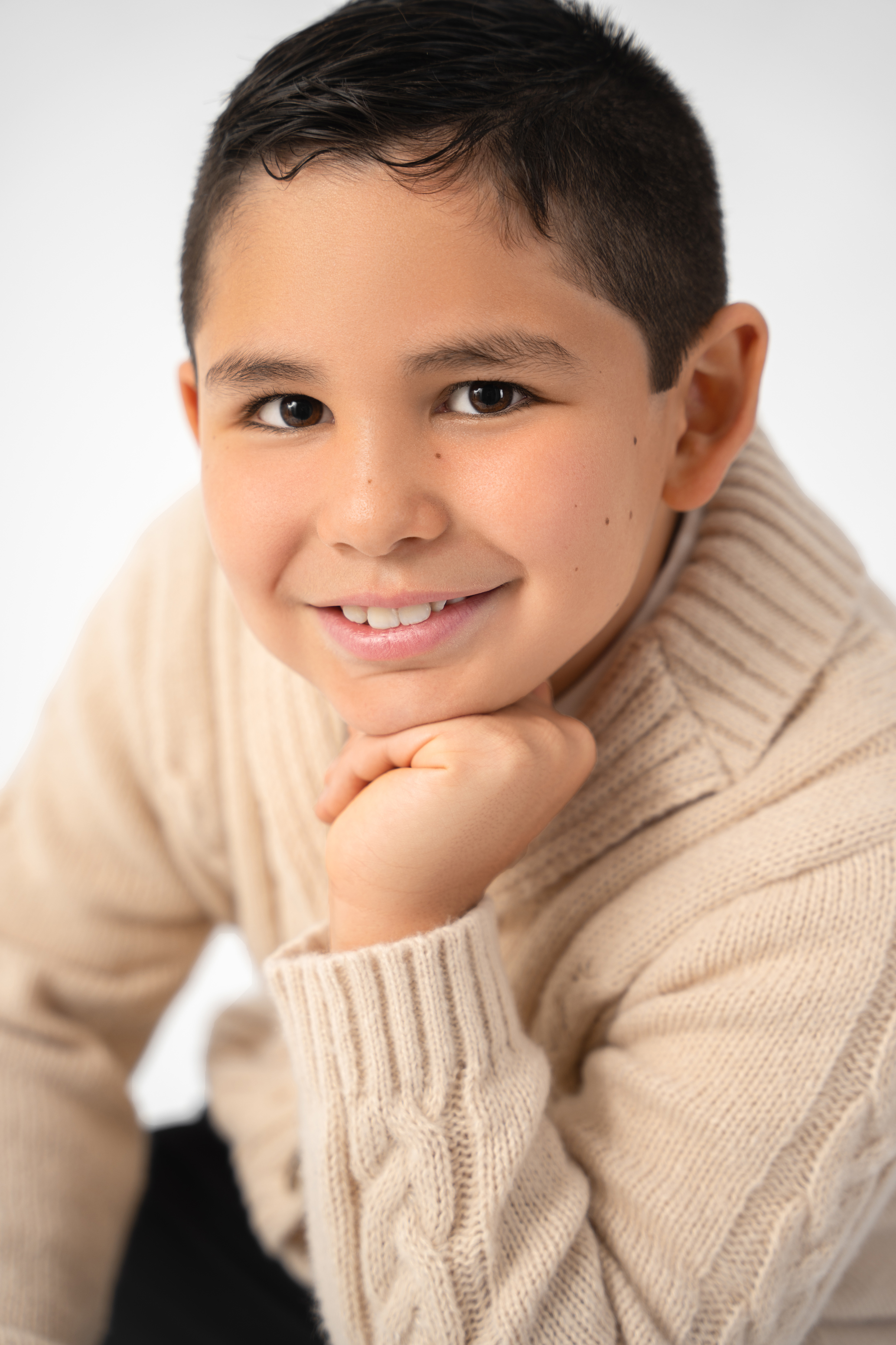 Close-up studio portrait of a smiling boy in a cream knit sweater against a soft white background