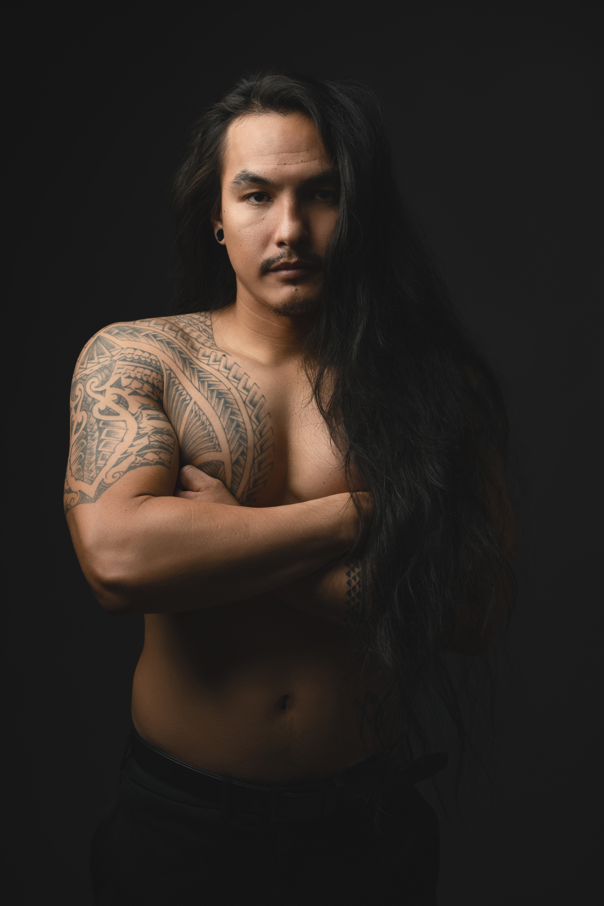 Low-key studio portrait of a long-haired man with tattoos posing shirtless against a dark background