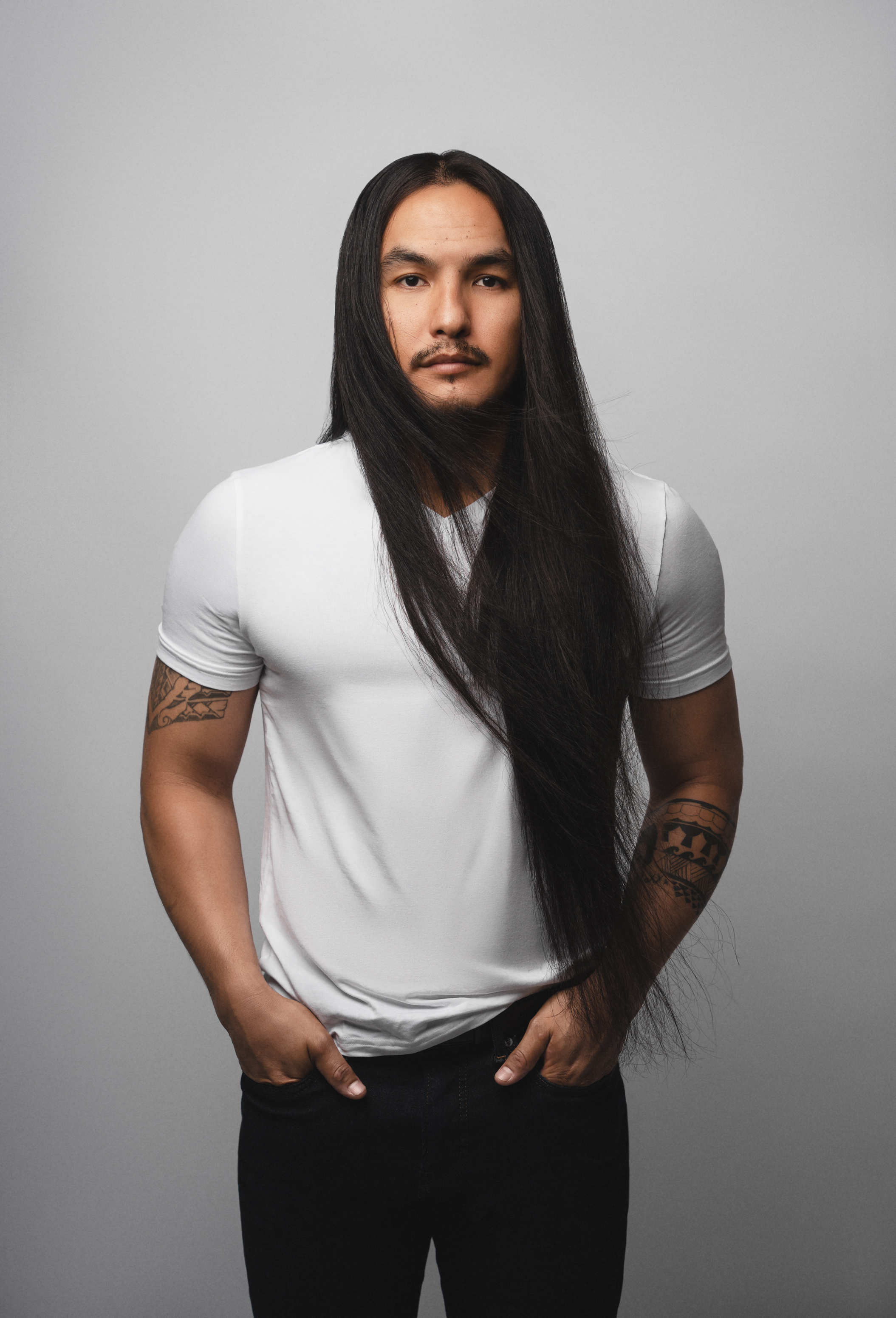 Full-length studio portrait of a long-haired man in a white T-shirt against a light gray background