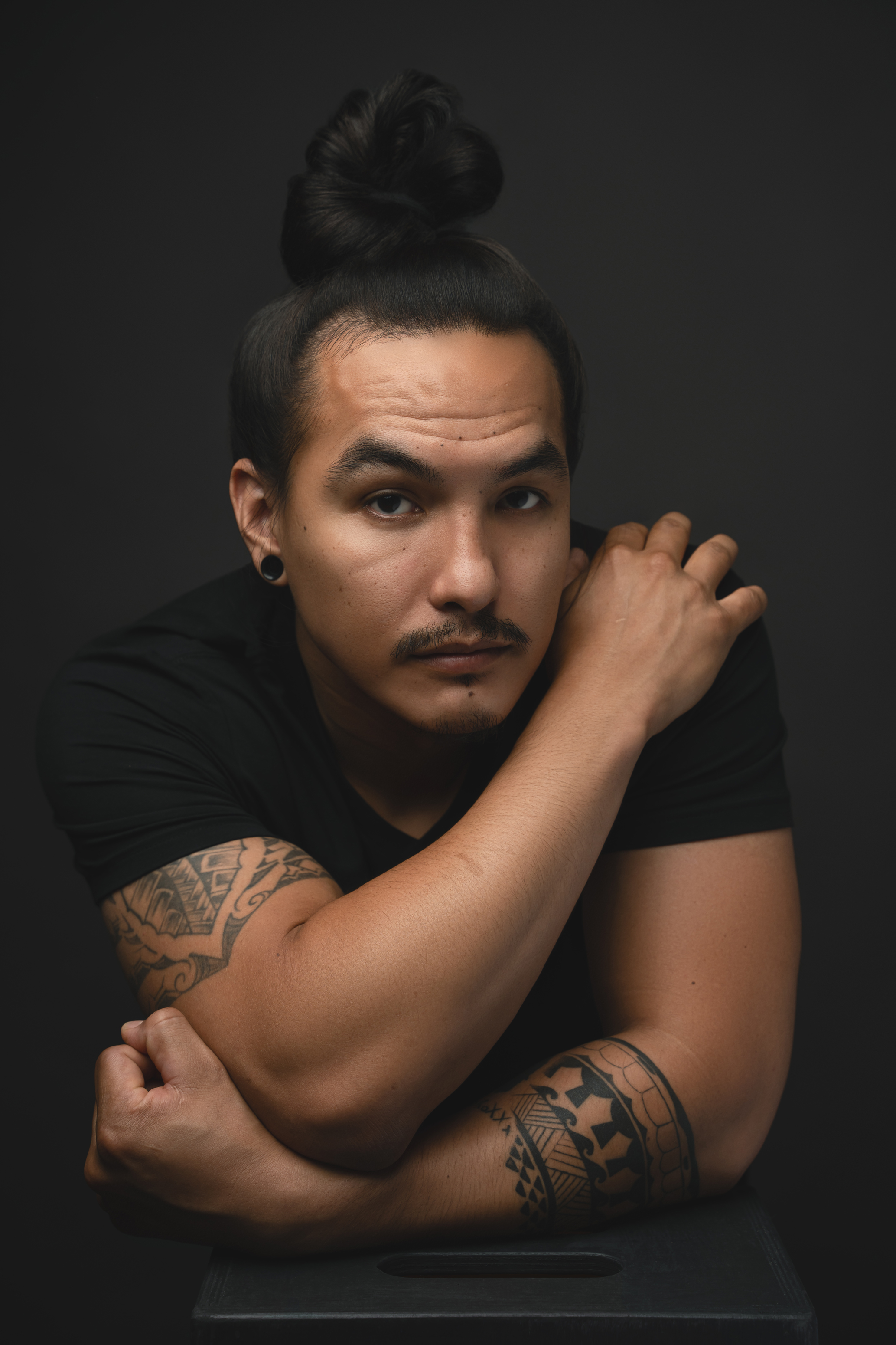 Moody seated studio portrait of a tattooed man with his hair tied up against a dark background