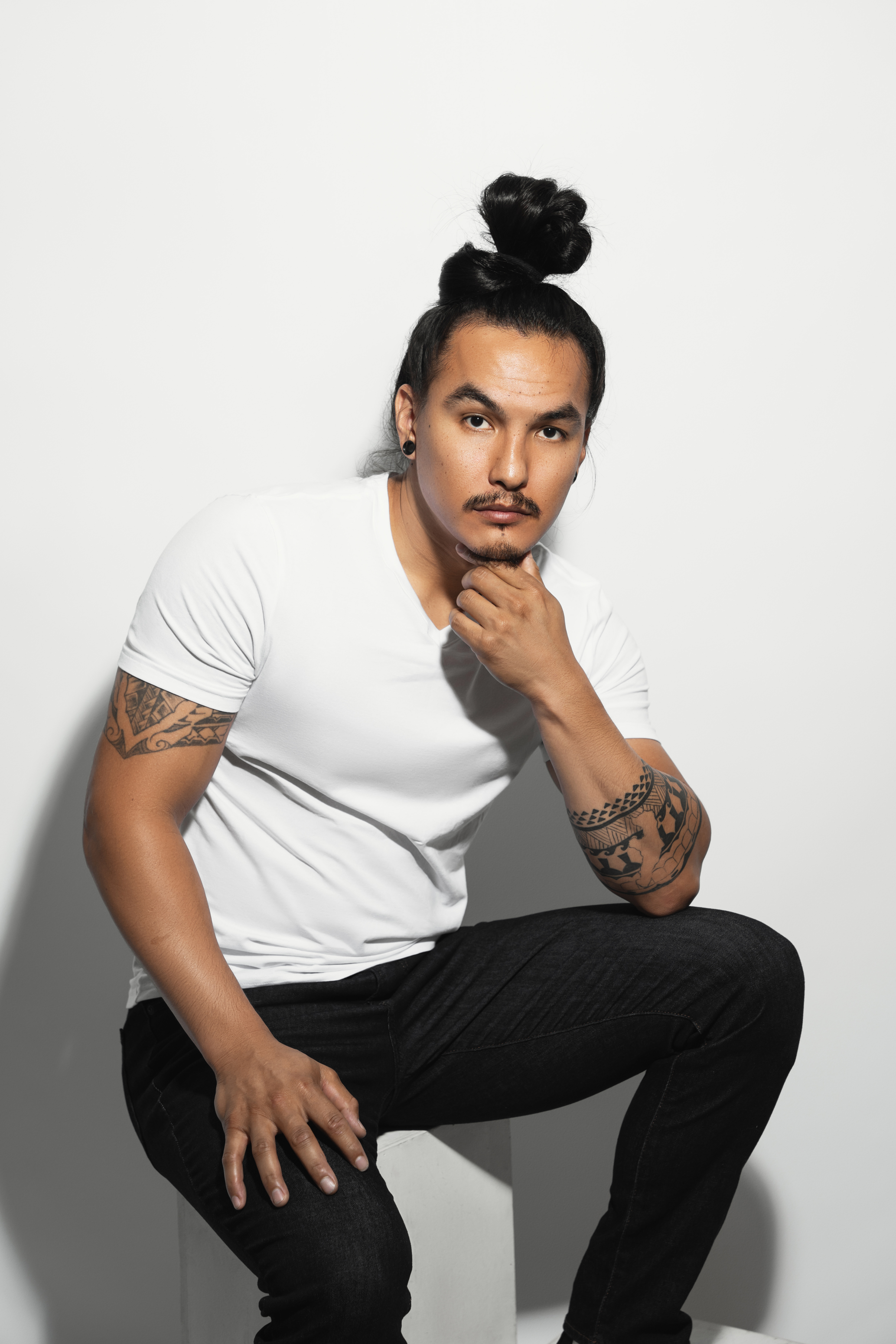 Seated studio portrait of a tattooed man in a white T-shirt posing against a bright neutral background