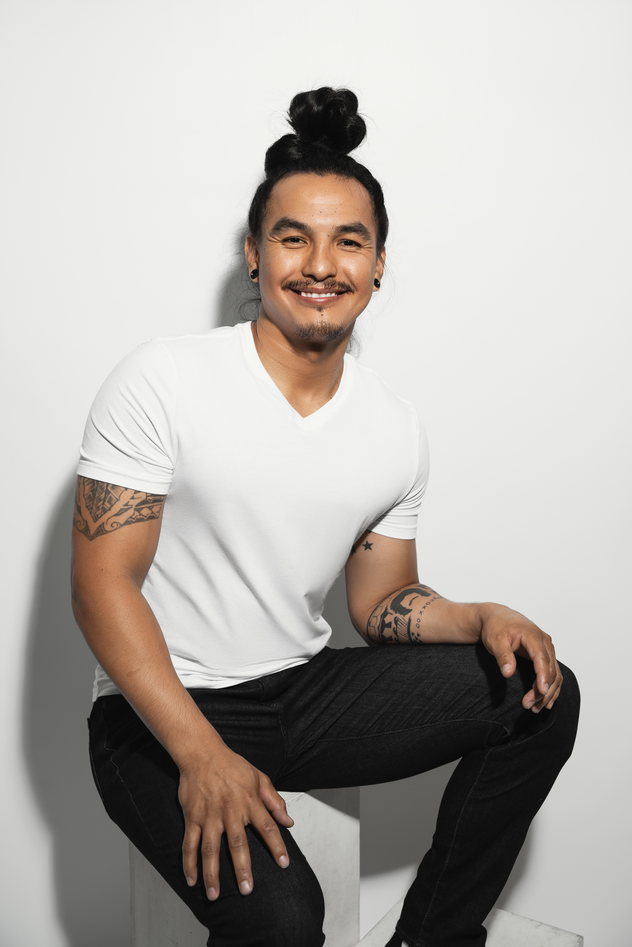Relaxed studio portrait of a smiling man with his hair tied up, seated against a light background