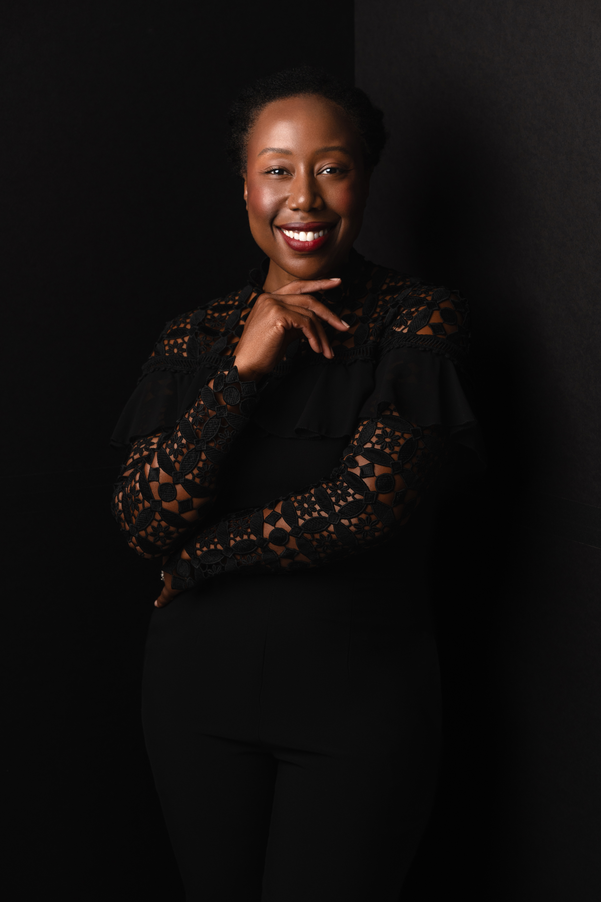 Low-key studio portrait of a smiling woman in a black lace top posing with one hand beneath her chin