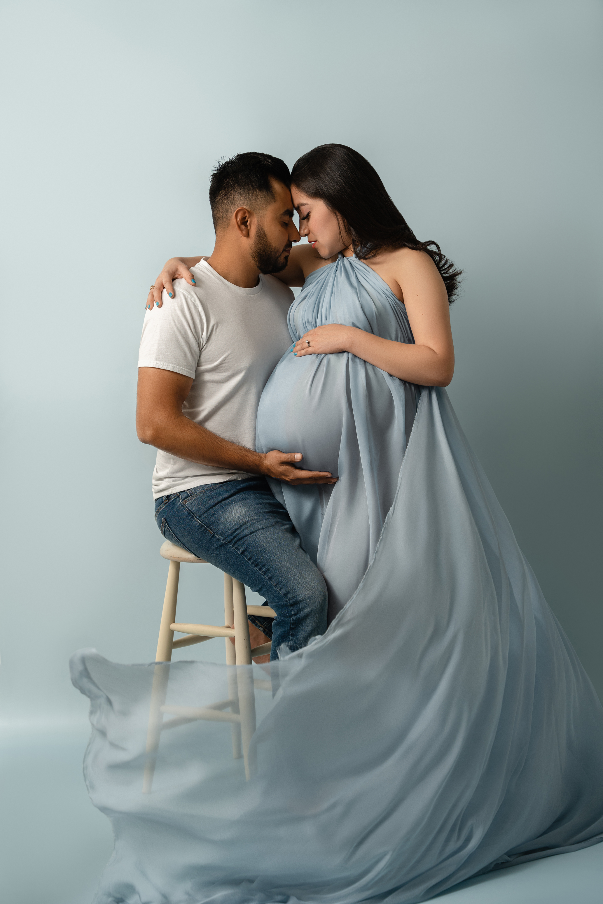 Studio maternity portrait of an expecting couple in a soft blue setup, seated closely together