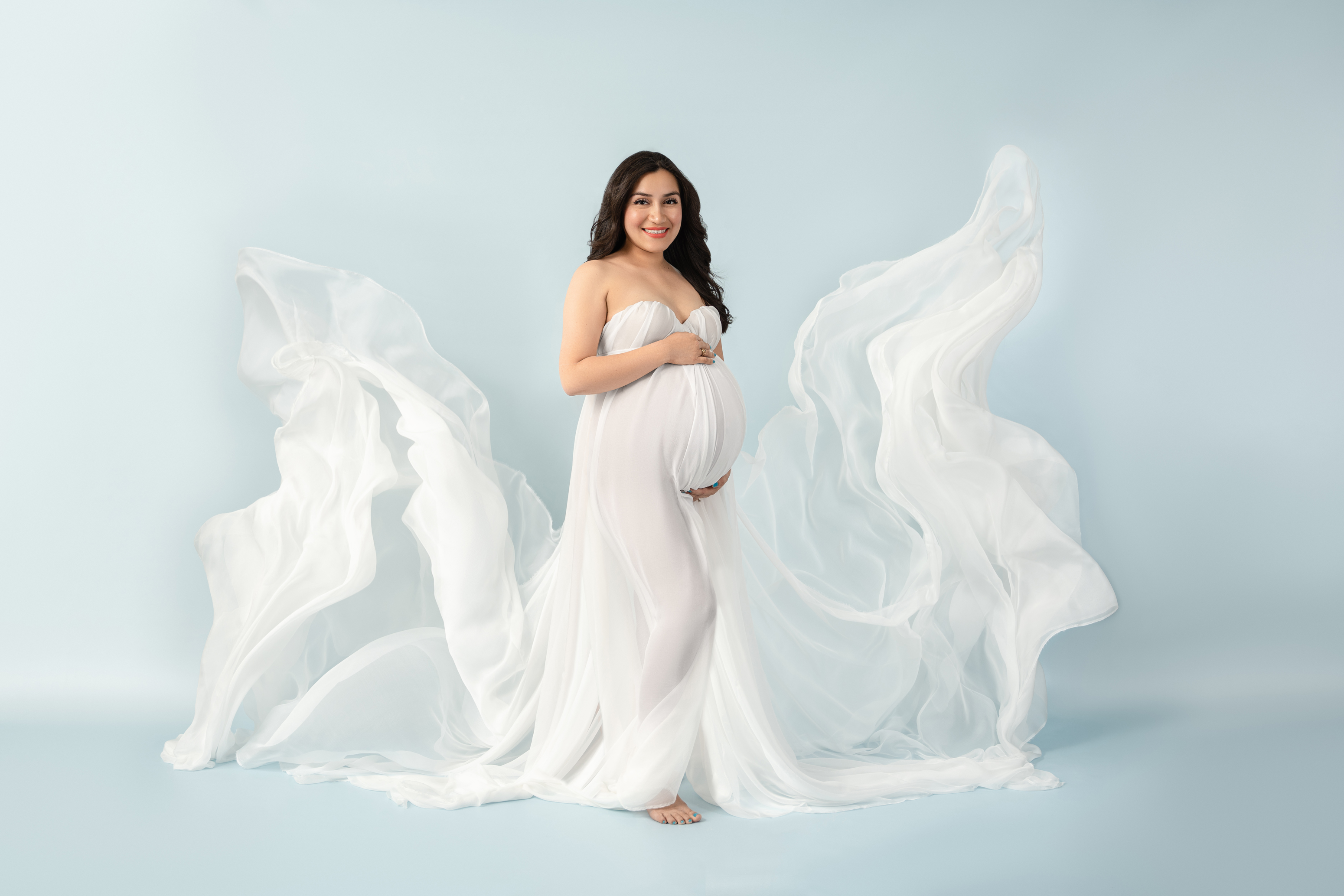 Wide maternity portrait of a woman in a flowing white gown with airy fabric against a pale blue studio background