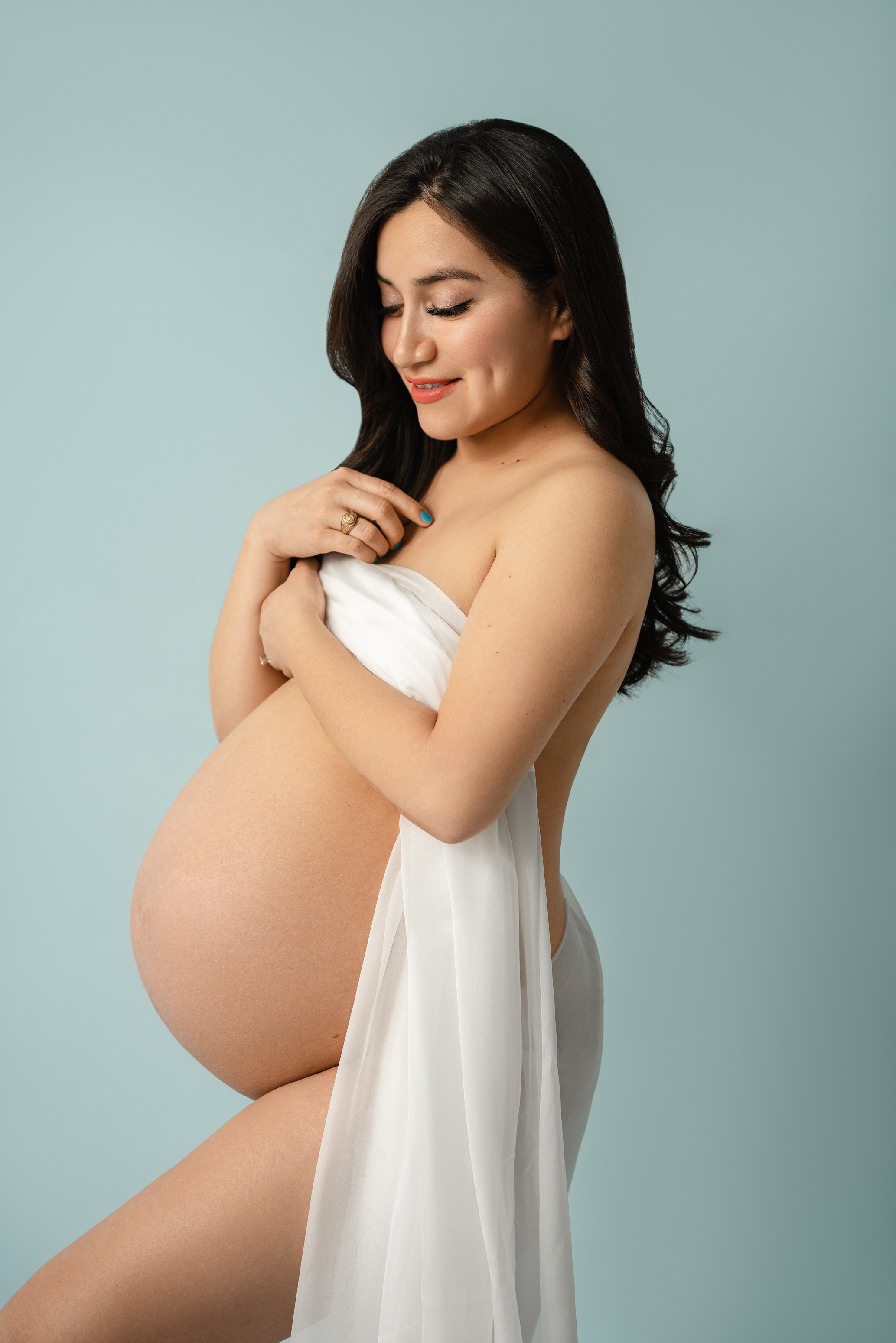 Minimal maternity portrait of a woman draped in white fabric against a soft blue background