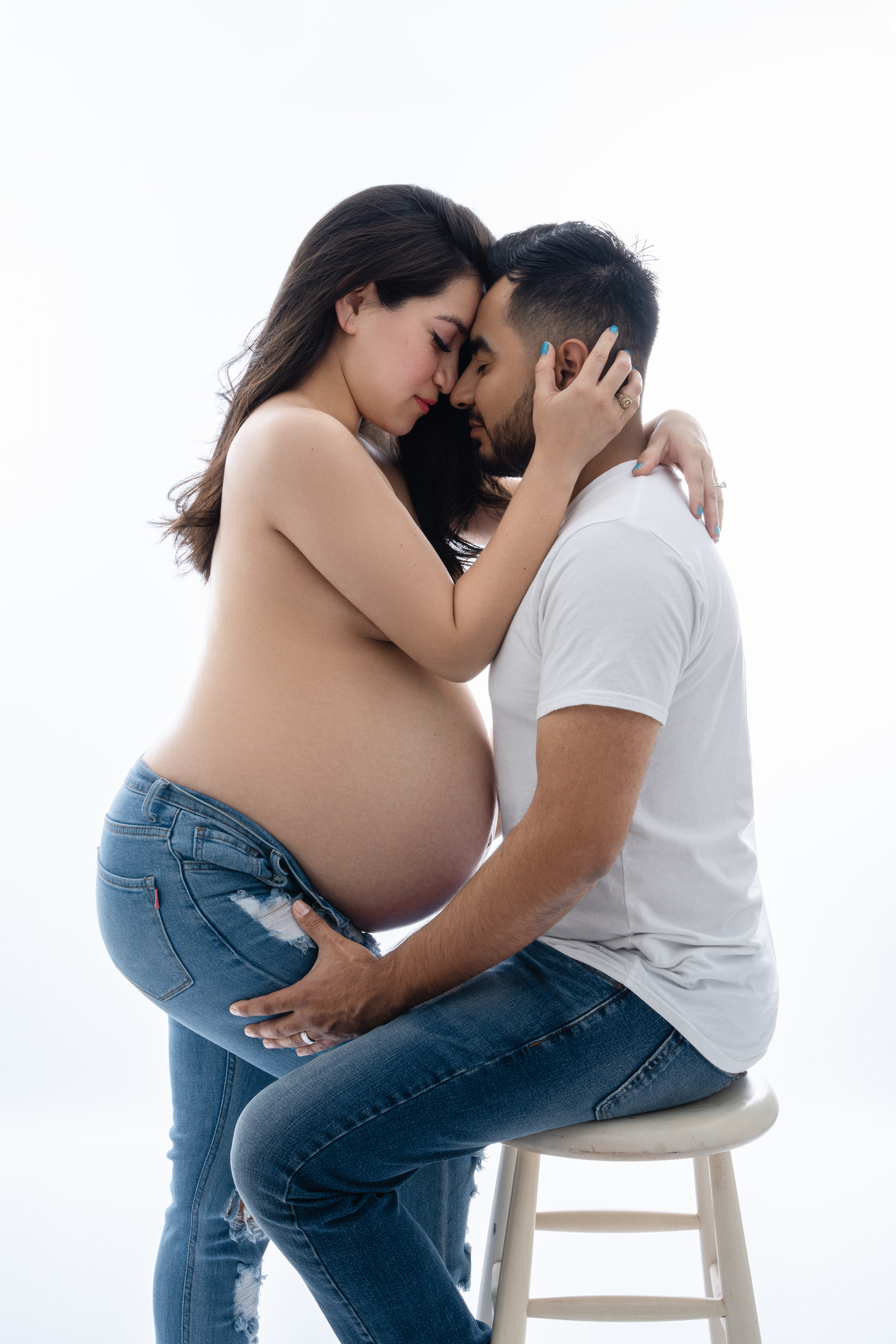 Intimate studio maternity portrait of an expecting couple seated together against a bright white background