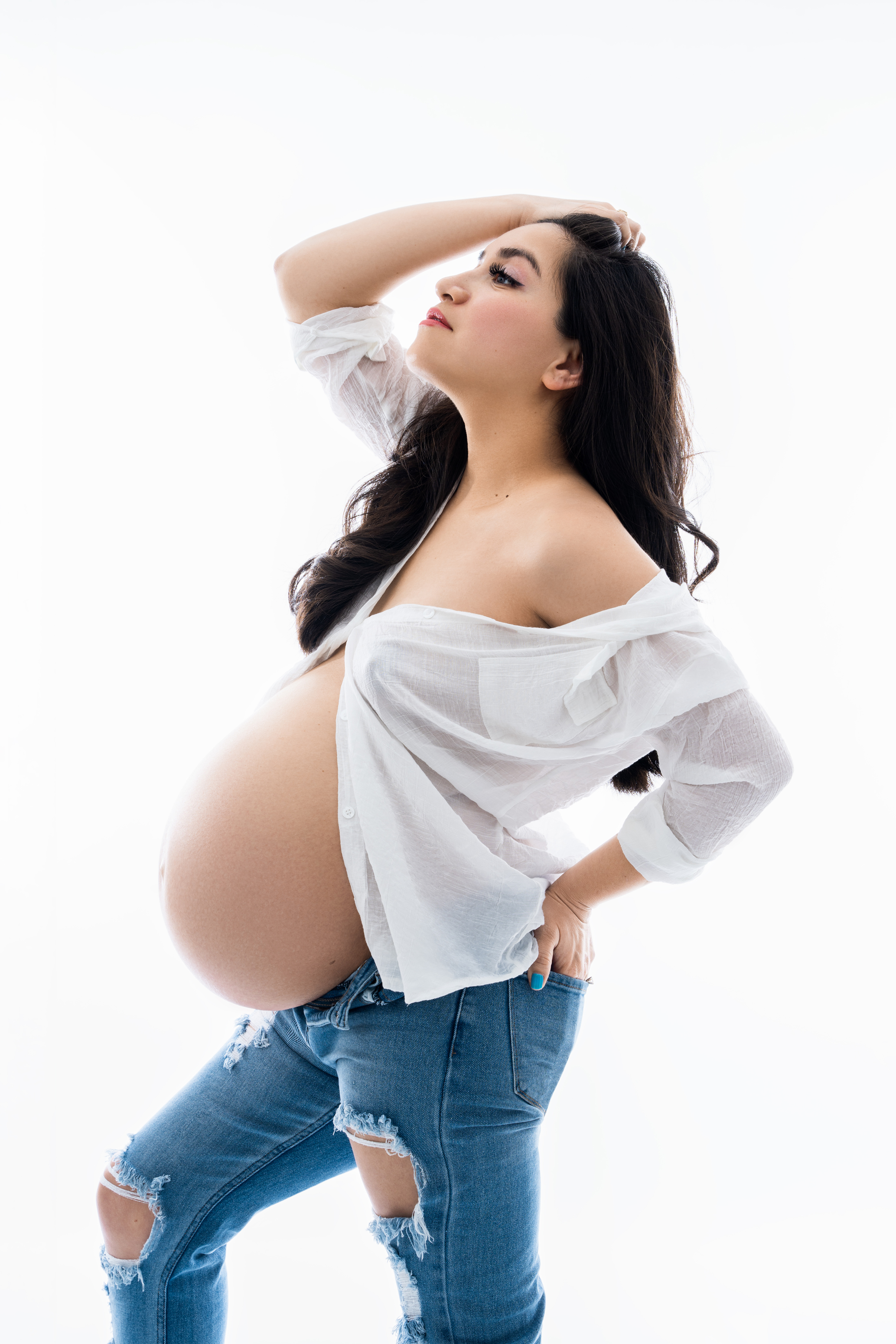 Bright studio maternity portrait of a woman in an open white shirt and jeans posing against a clean white background