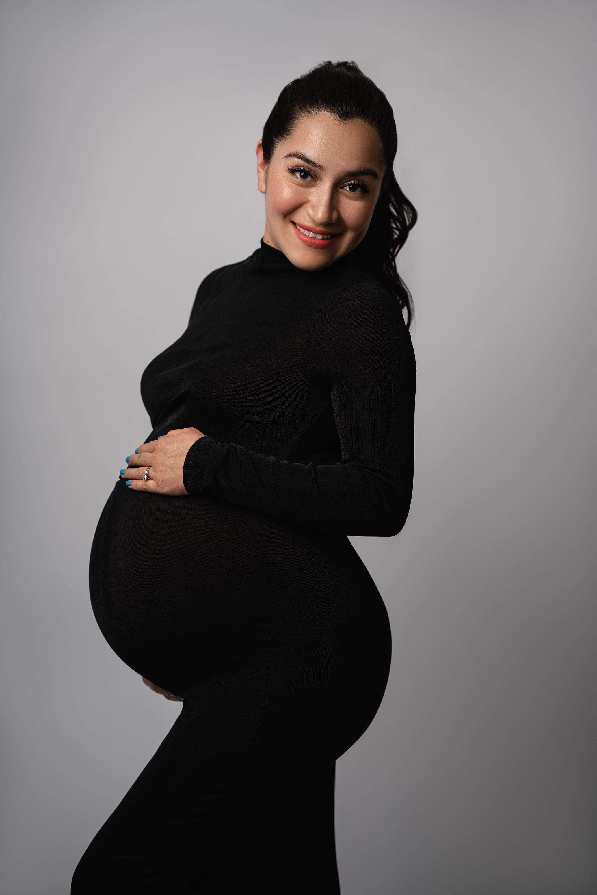 Studio maternity portrait of a woman in a fitted black dress against a soft gray background