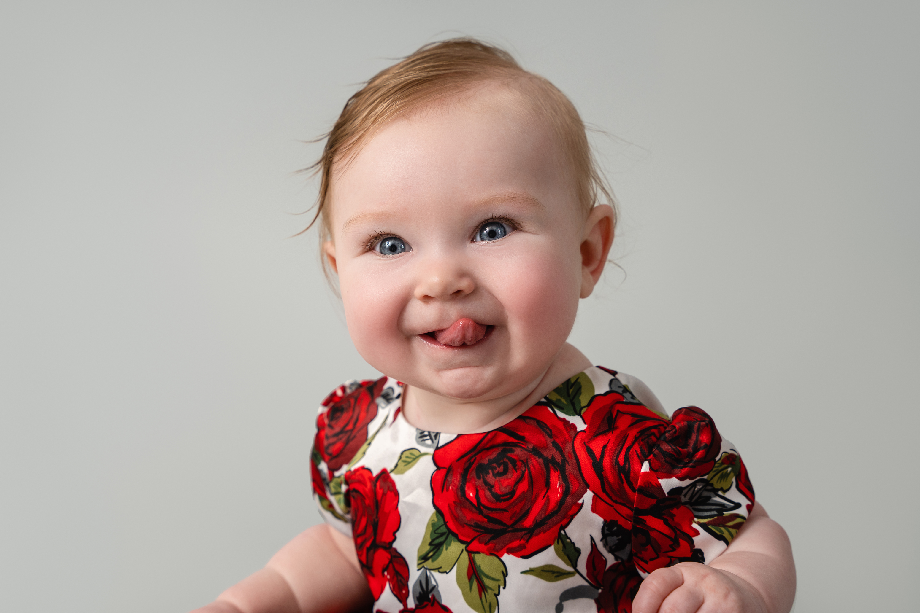 Close-up studio portrait of a smiling baby in a red floral dress against a soft gray background