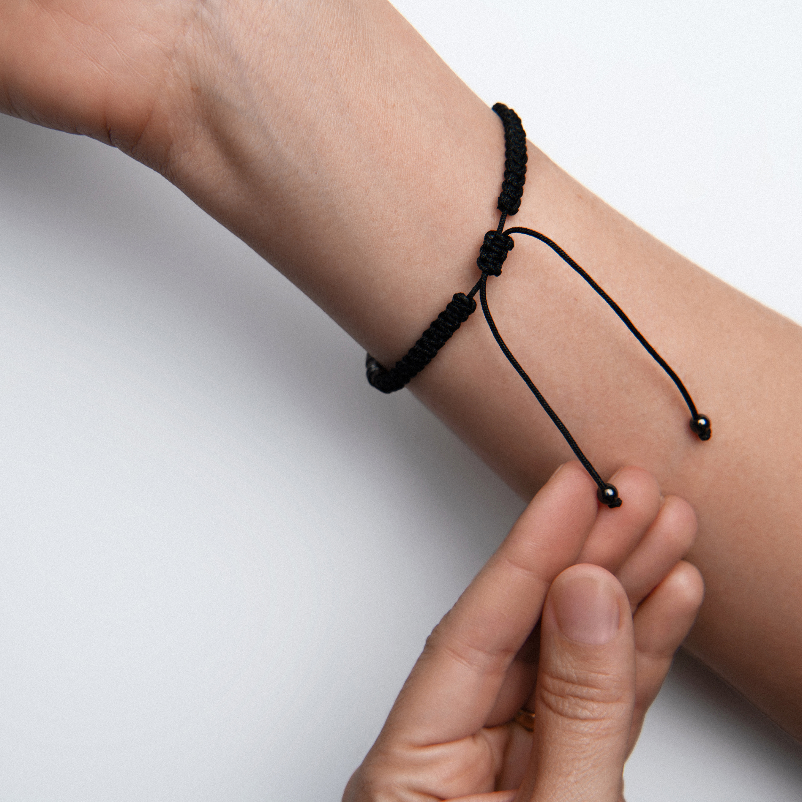 Close-up product image of a black cord bracelet being adjusted on the wrist against a clean neutral background