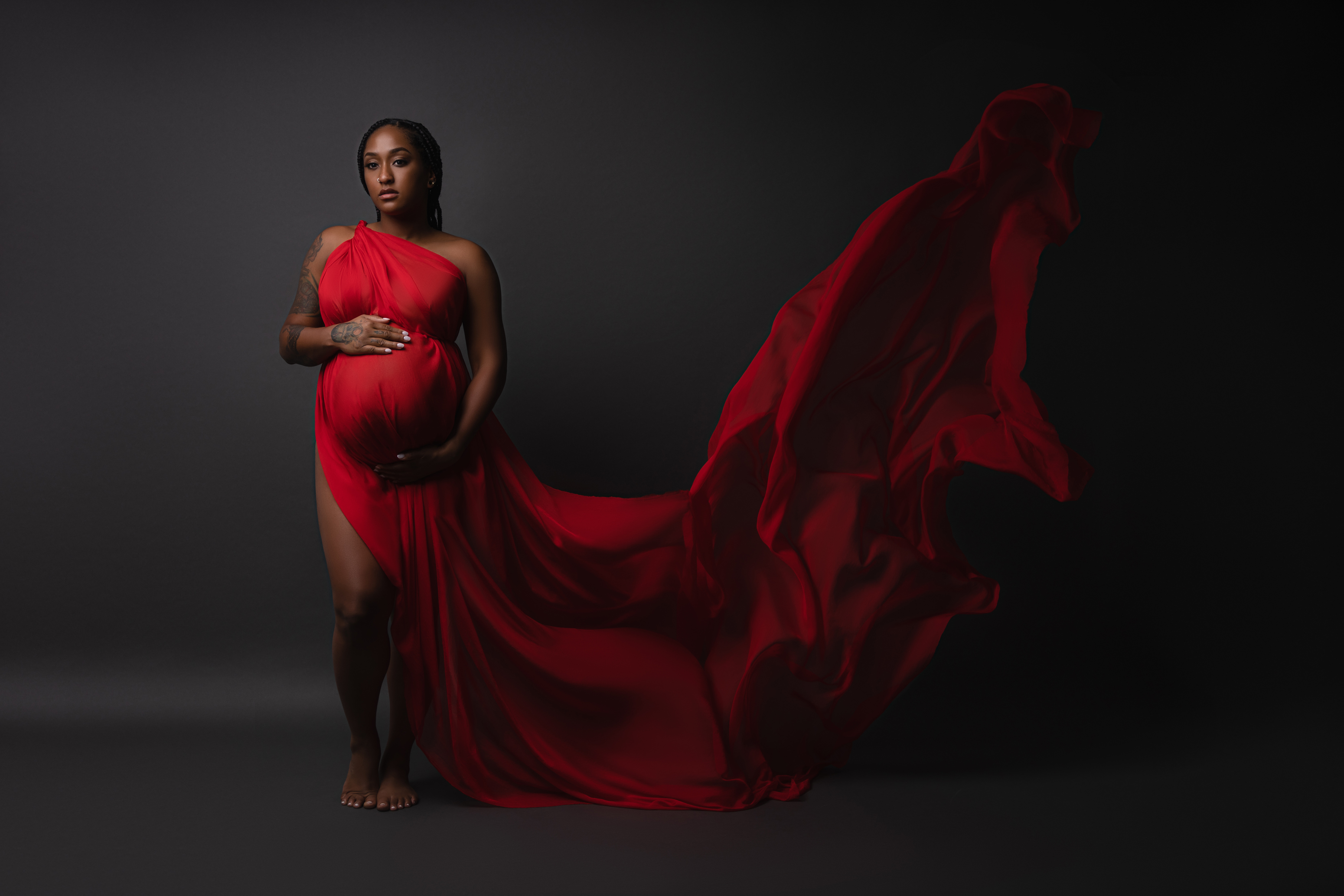 Dramatic maternity portrait in a flowing red gown with fabric lifted through a dark studio scene