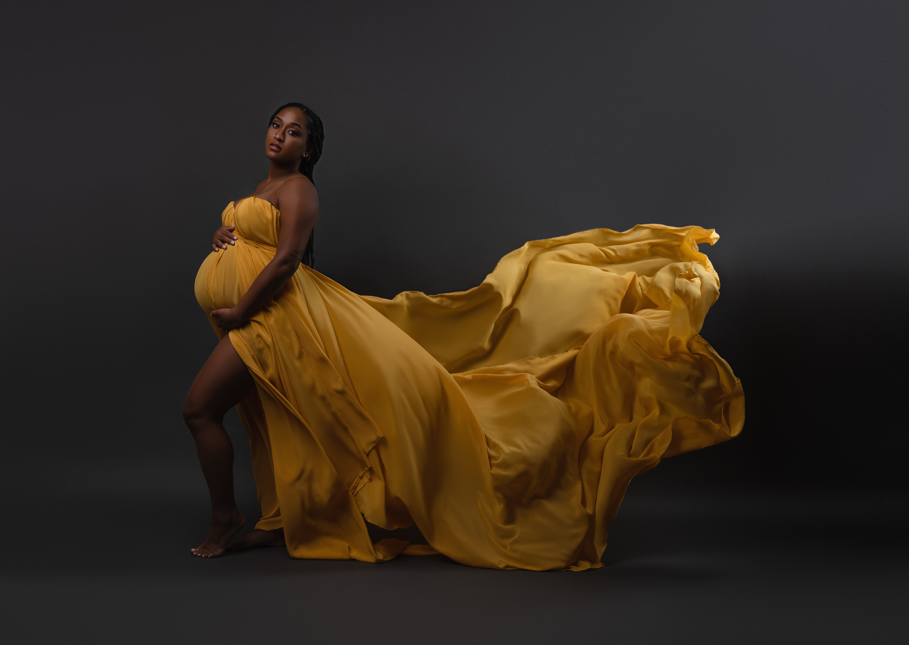 Full-length maternity portrait in a flowing golden gown with fabric sweeping across a dark studio setting