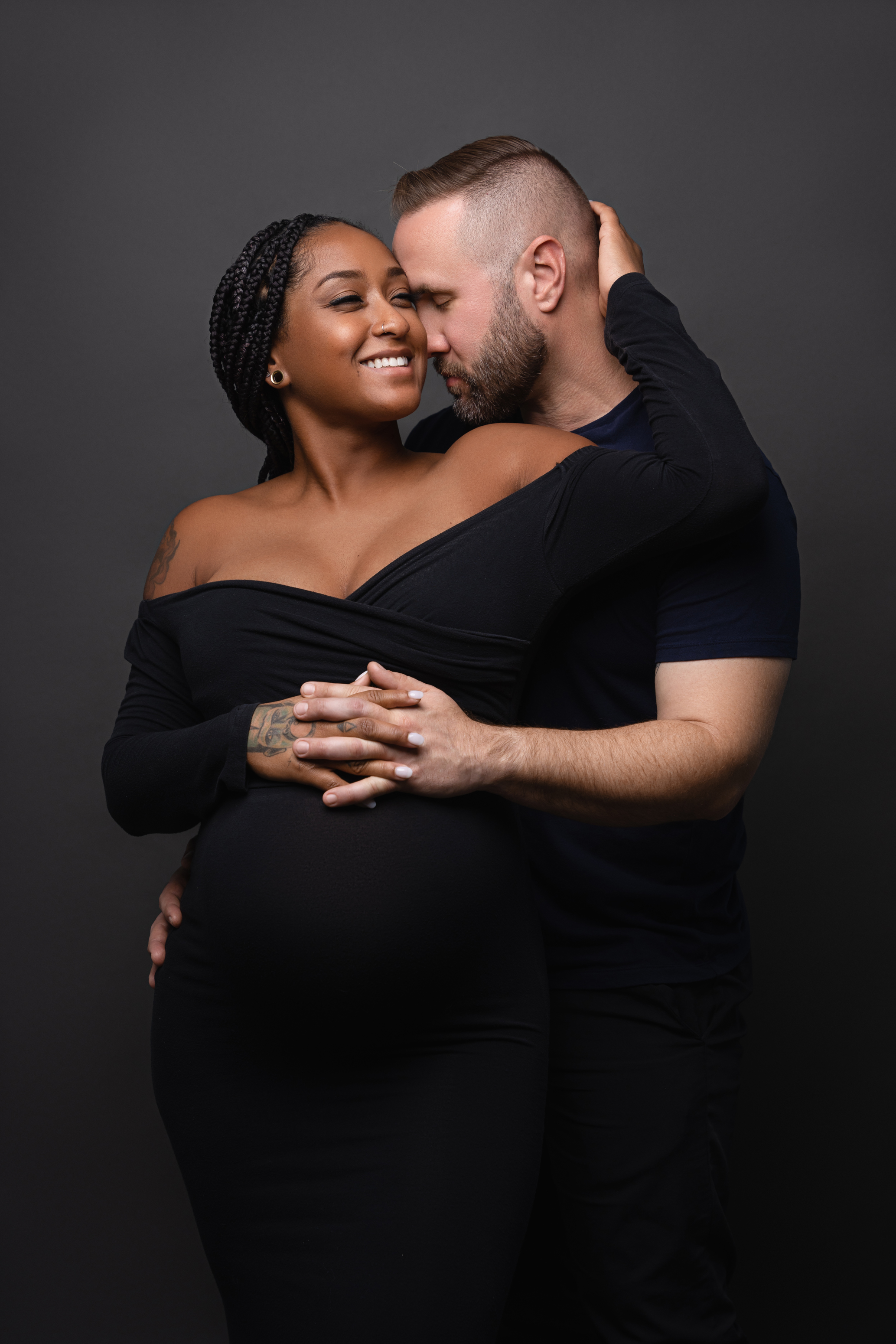 Intimate studio maternity portrait of an expecting couple embracing against a dark neutral background