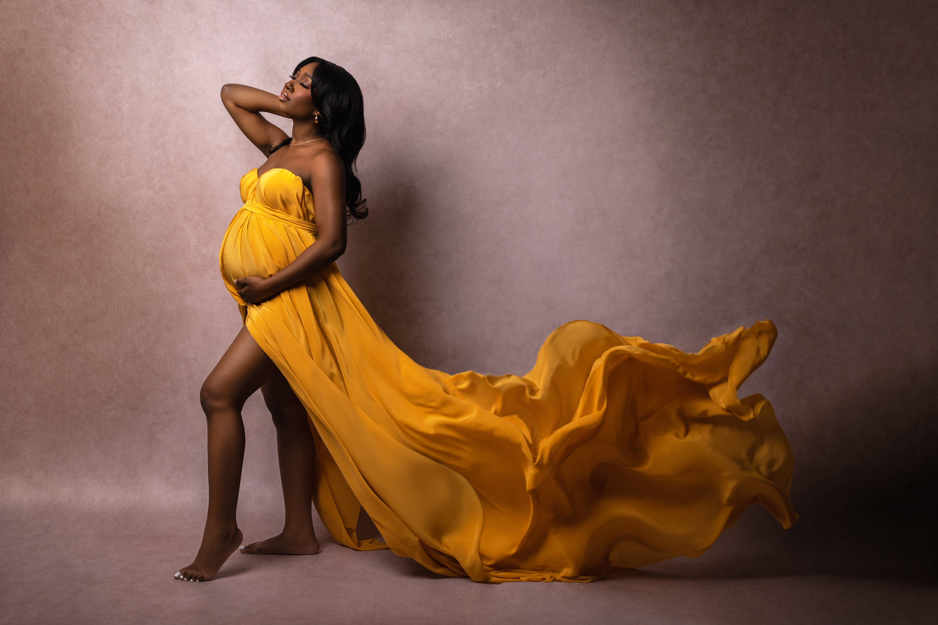 Dramatic maternity portrait in a flowing golden gown with fabric sweeping through a dark neutral studio setting