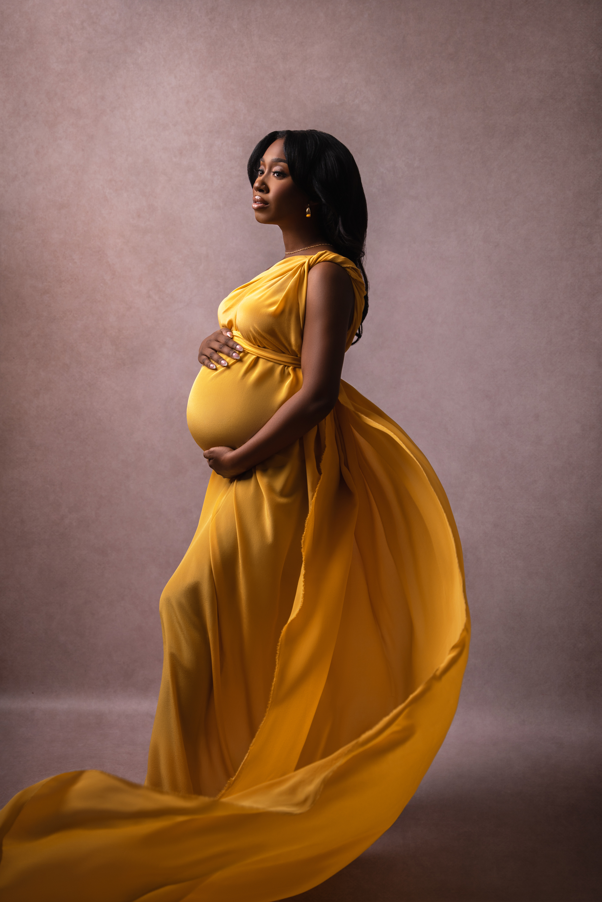 Side-profile maternity portrait in a flowing golden gown against a softly textured neutral background