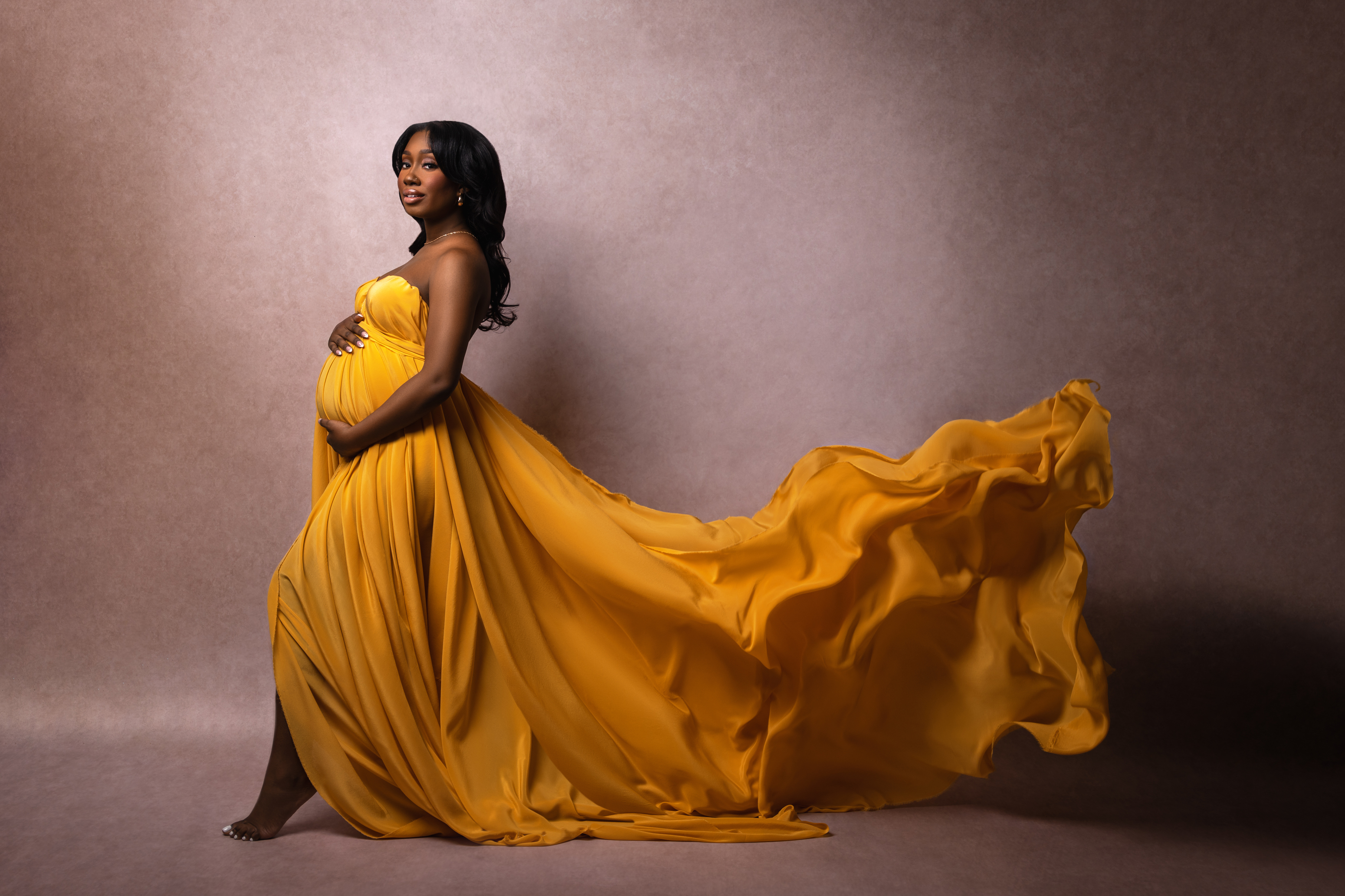 Wide maternity portrait in a golden gown with flowing fabric extending across the studio floor