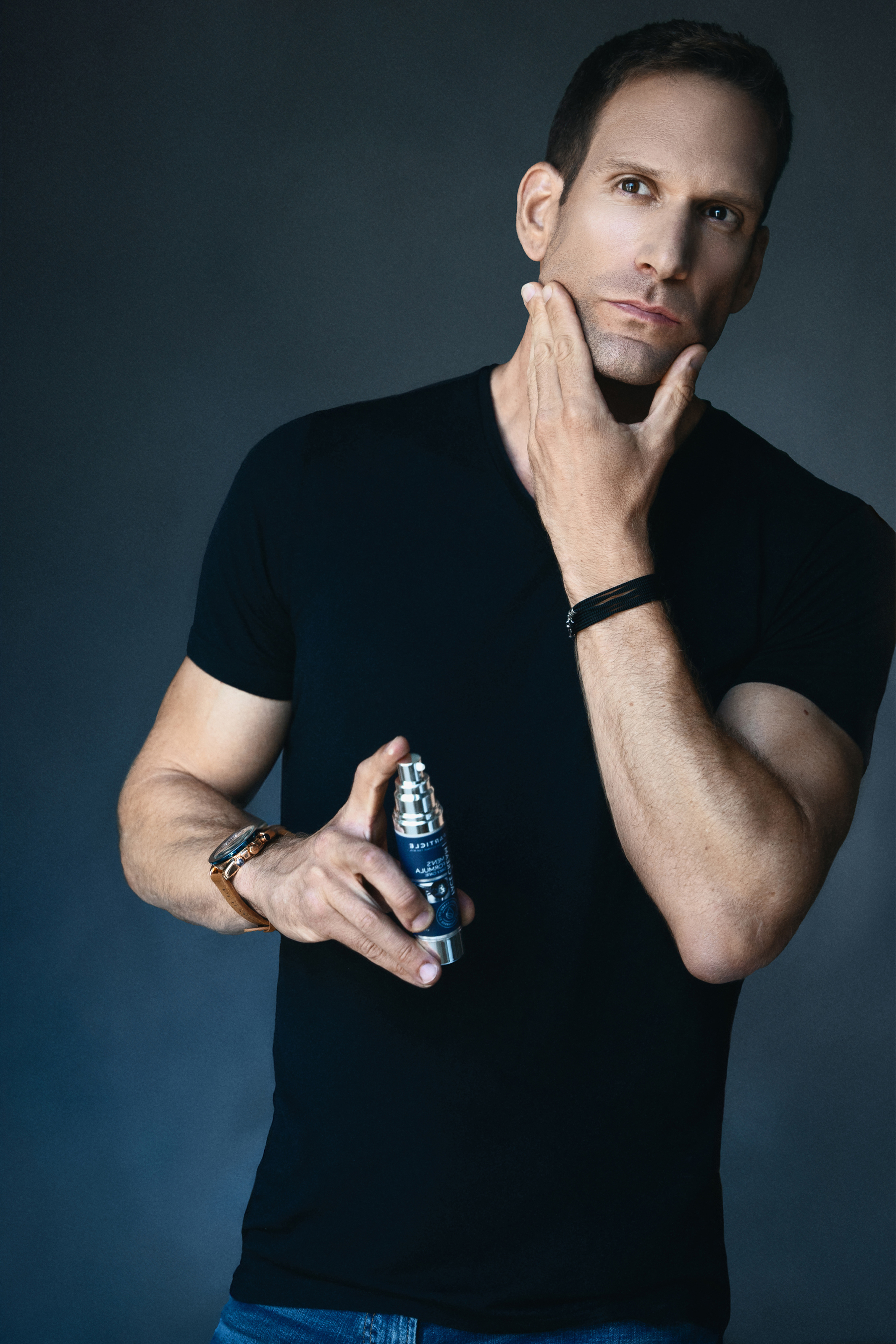 Commercial portrait of a man in a black T-shirt holding a men’s skincare product against a dark blue background