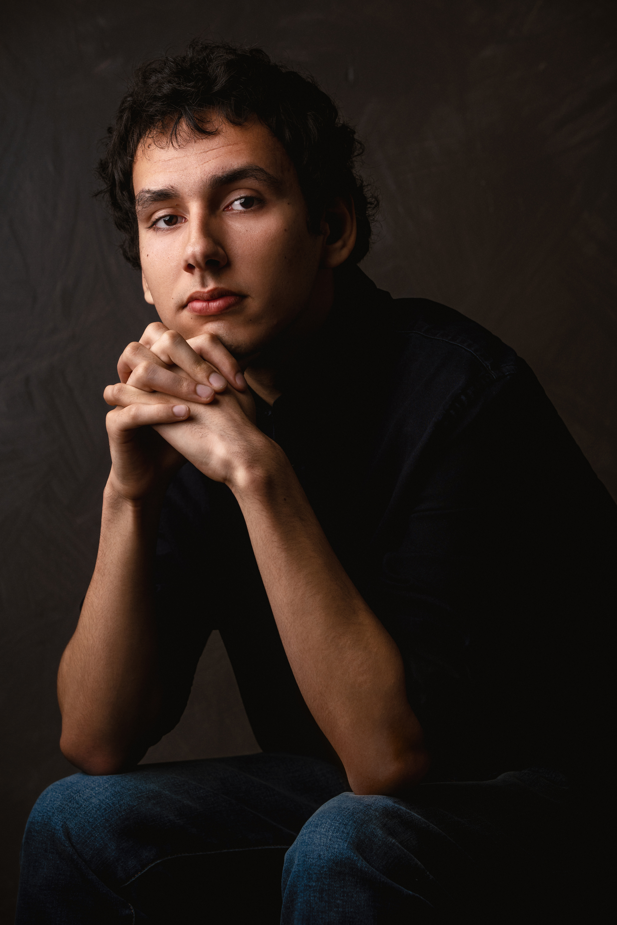 Low-key seated portrait of a man in dark clothing with warm directional light against a textured background