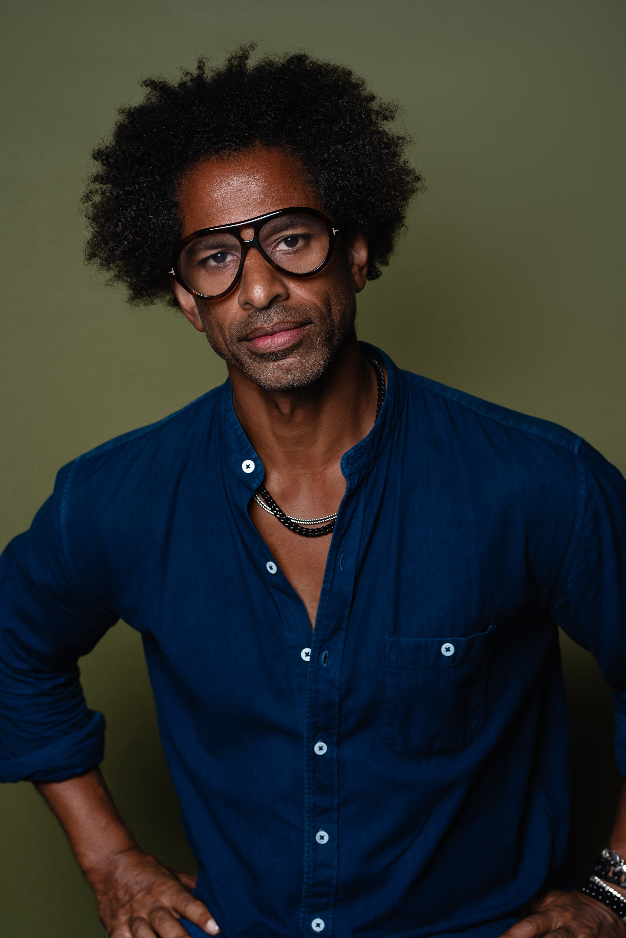 Studio portrait of a man in a dark blue shirt and black glasses against a muted olive background