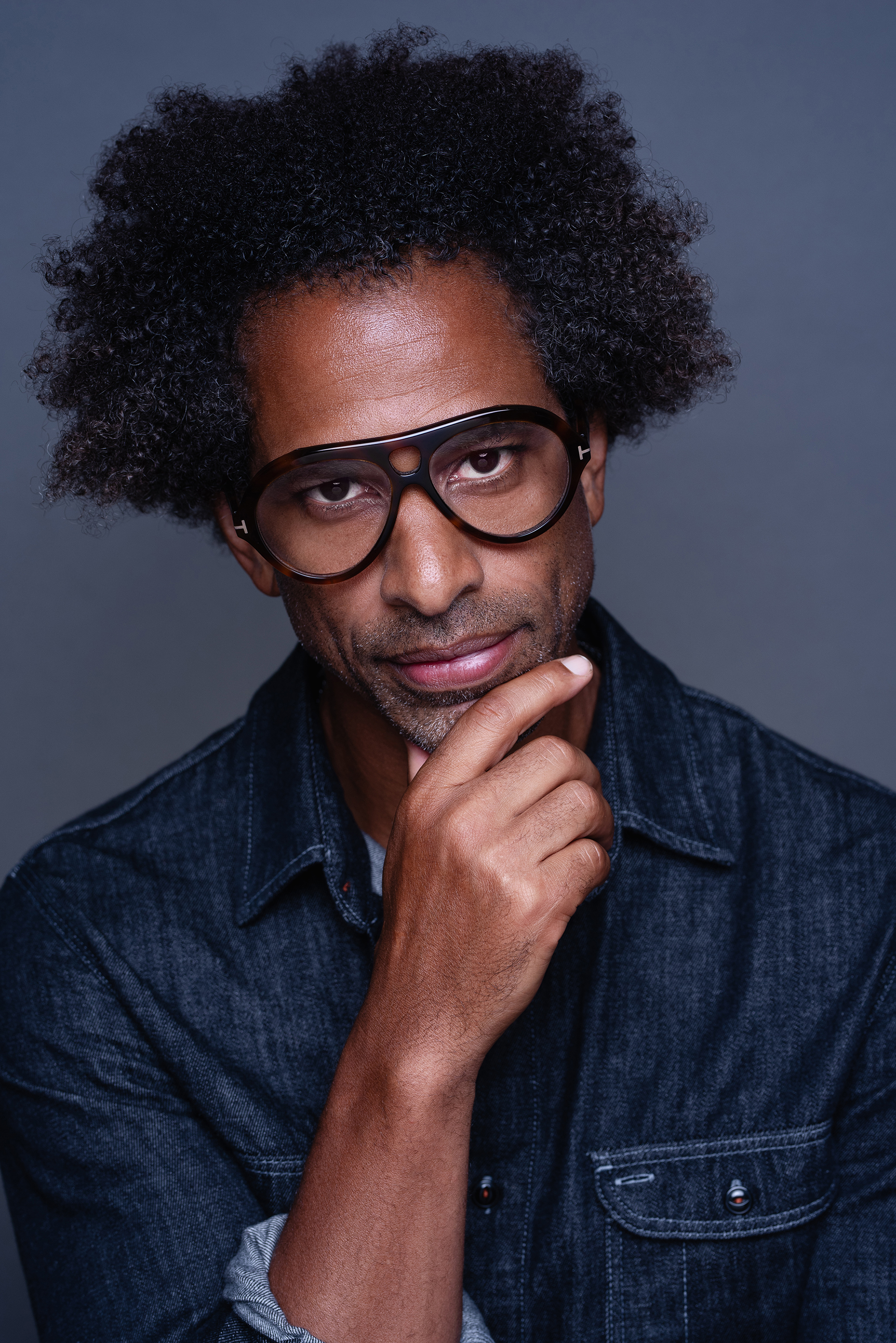 Personal branding portrait of a man in a dark denim shirt and black glasses posing with one hand beneath his chin