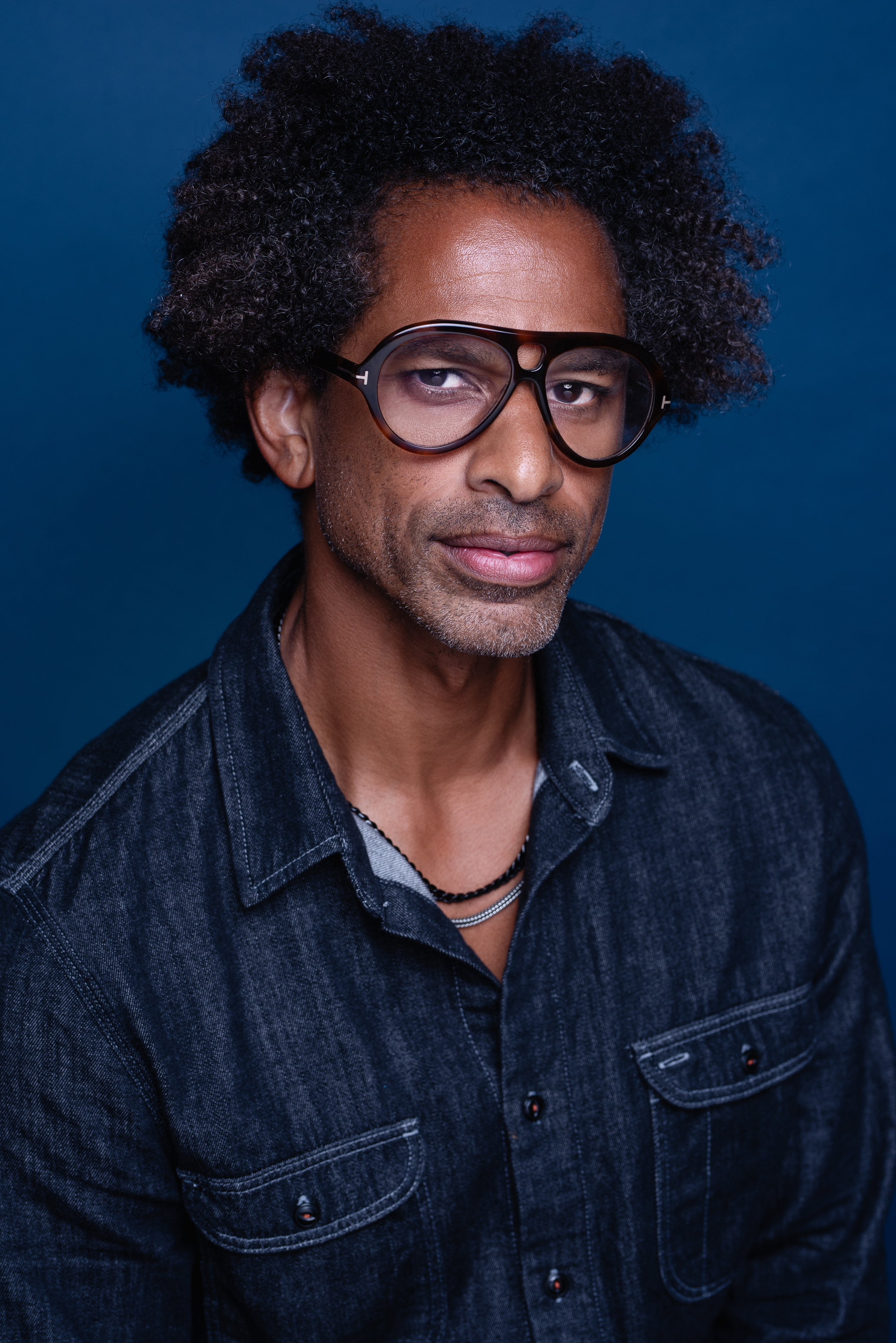 Studio portrait of a man in a dark denim shirt and oversized glasses against a rich blue background
