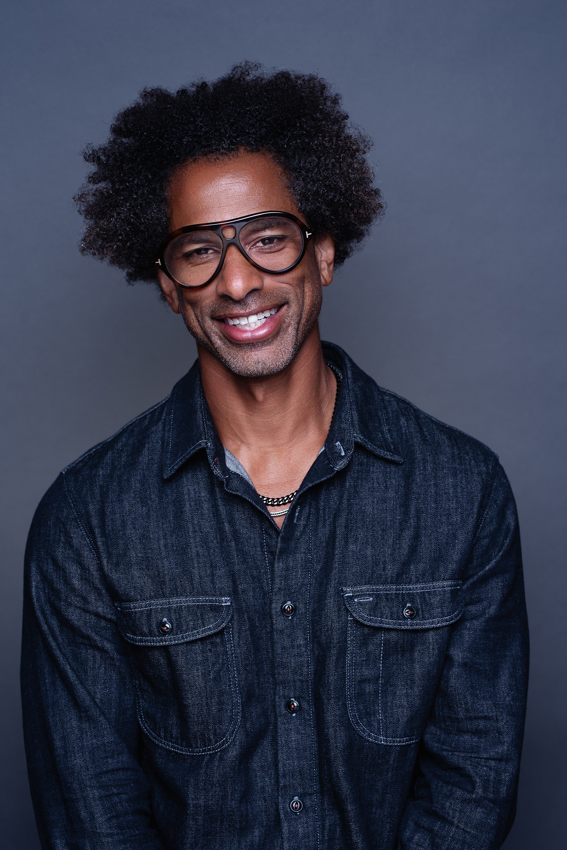 Smiling studio portrait of a man in a dark denim shirt and black glasses against a soft gray background