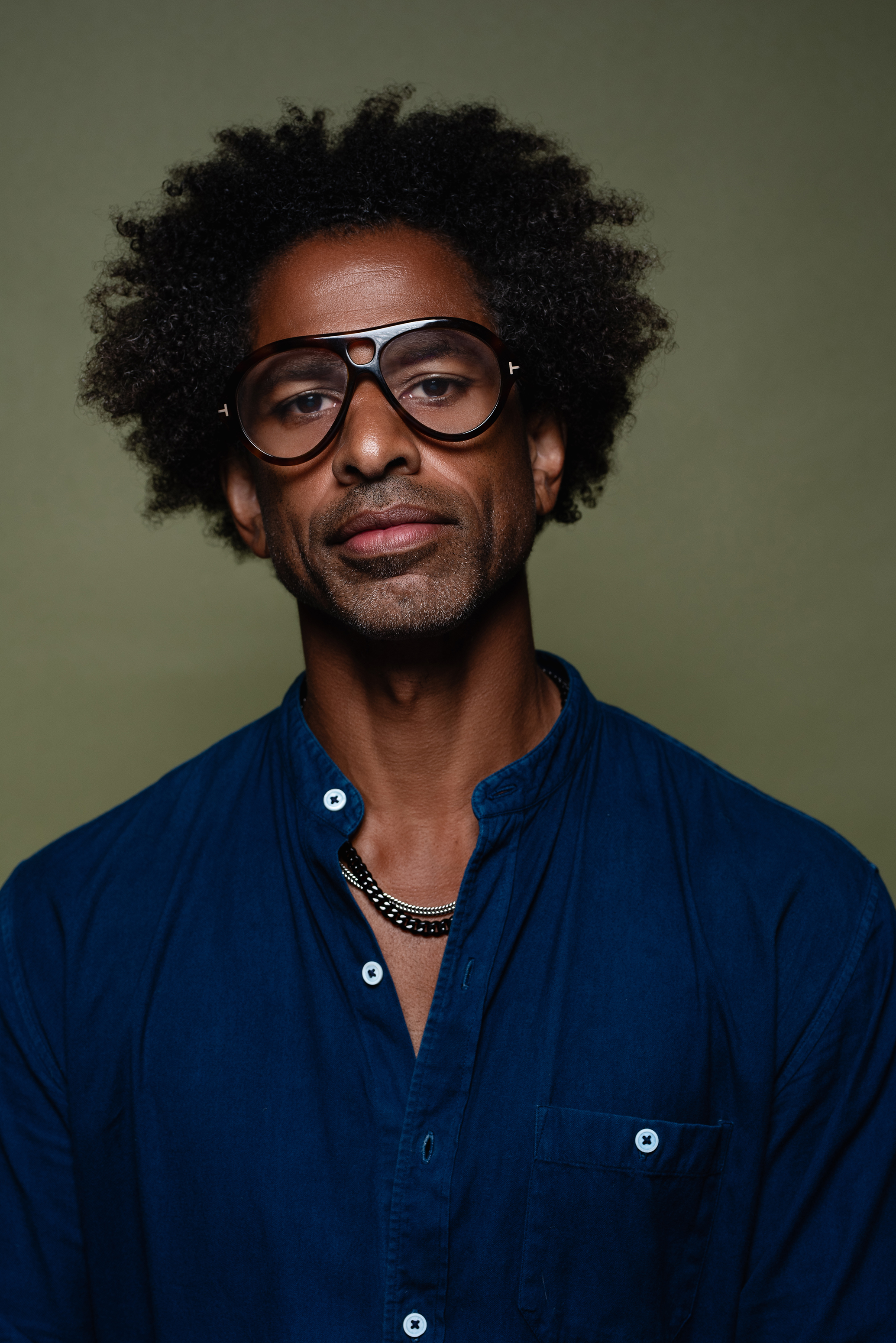 Close studio portrait of a man in a blue shirt and oversized glasses against a muted olive background