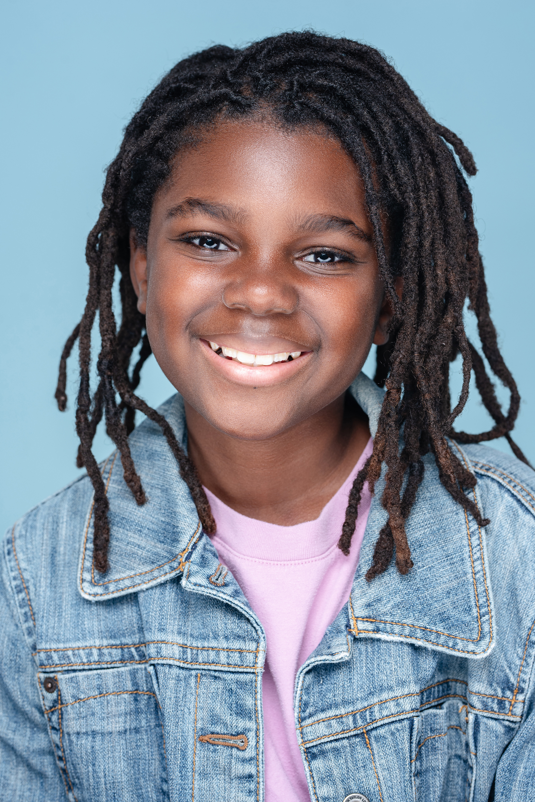 Smiling studio portrait of a child with locs in a denim jacket against a soft blue background