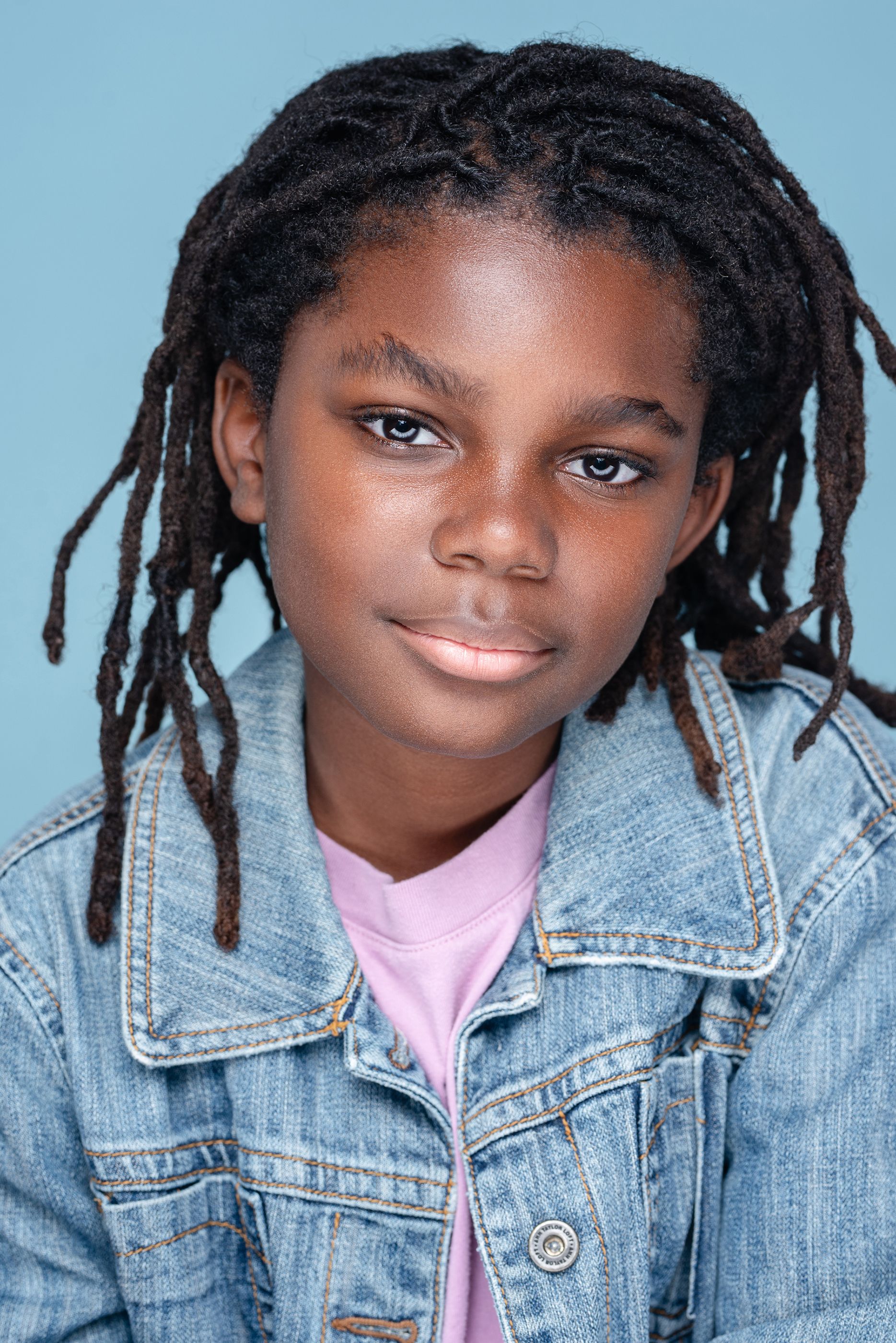 Close studio portrait of a child with locs in a denim jacket against a light blue background