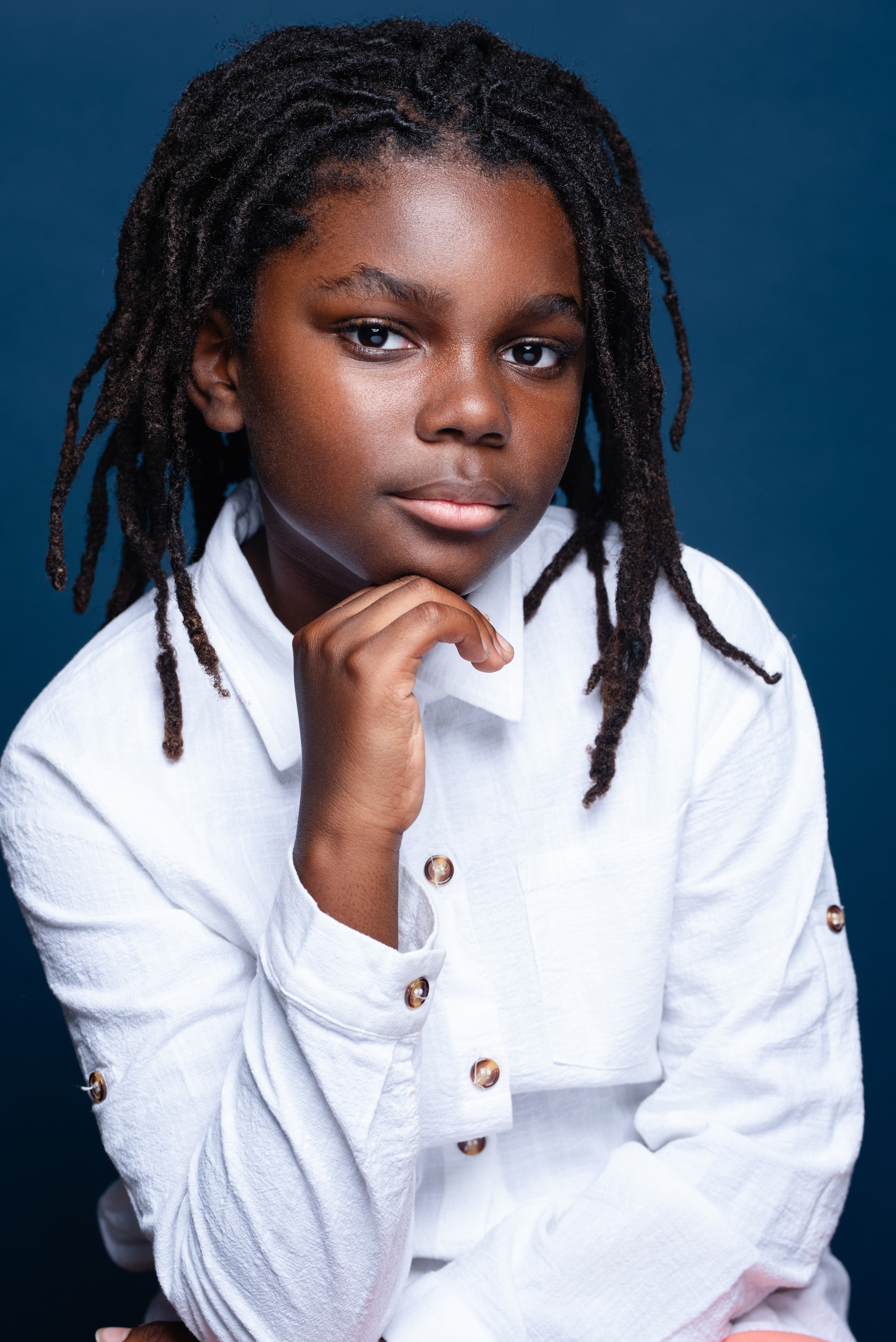Studio portrait of a child with locs in a white shirt posing against a deep blue background