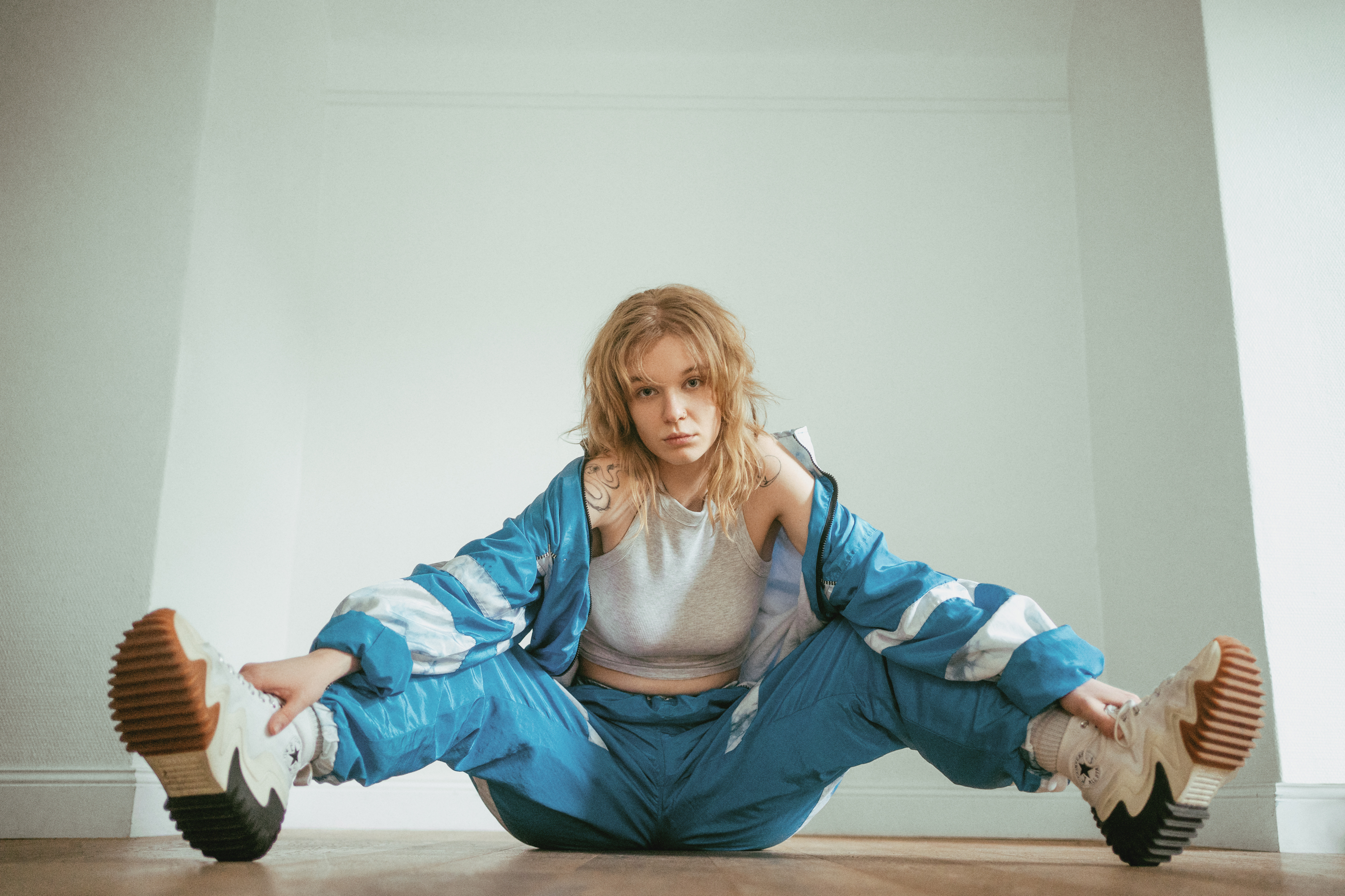 Editorial portrait of a woman in a blue tracksuit seated on the floor in a bright minimal interior