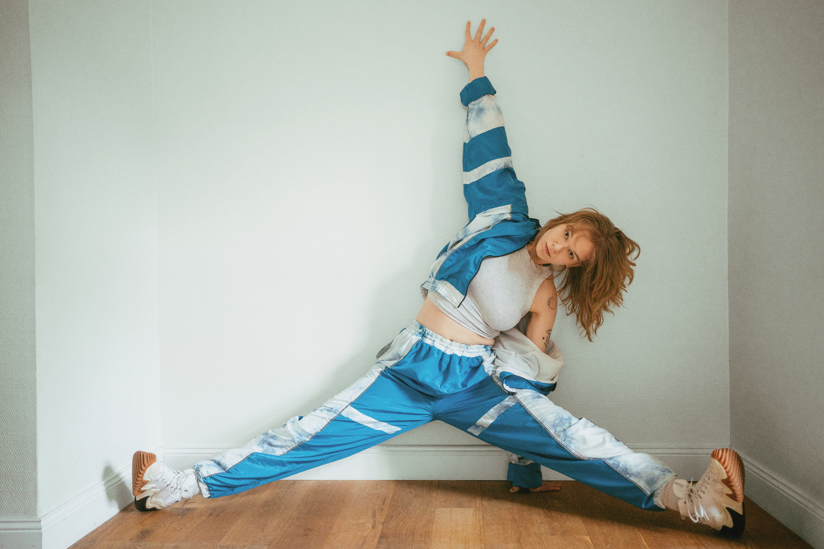 Dynamic editorial portrait of a woman in a blue tracksuit stretching across a bright interior space
