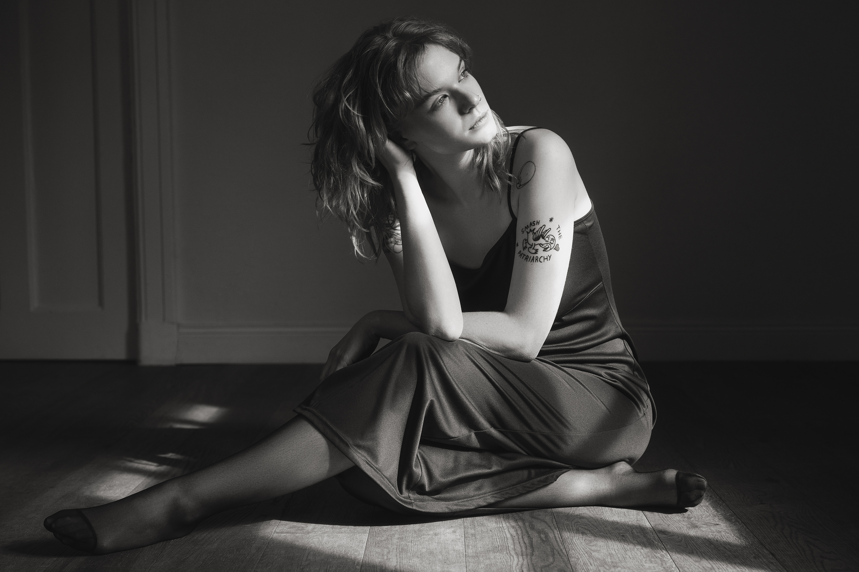 Black-and-white portrait of a woman seated on the floor in a dark dress with dramatic window light