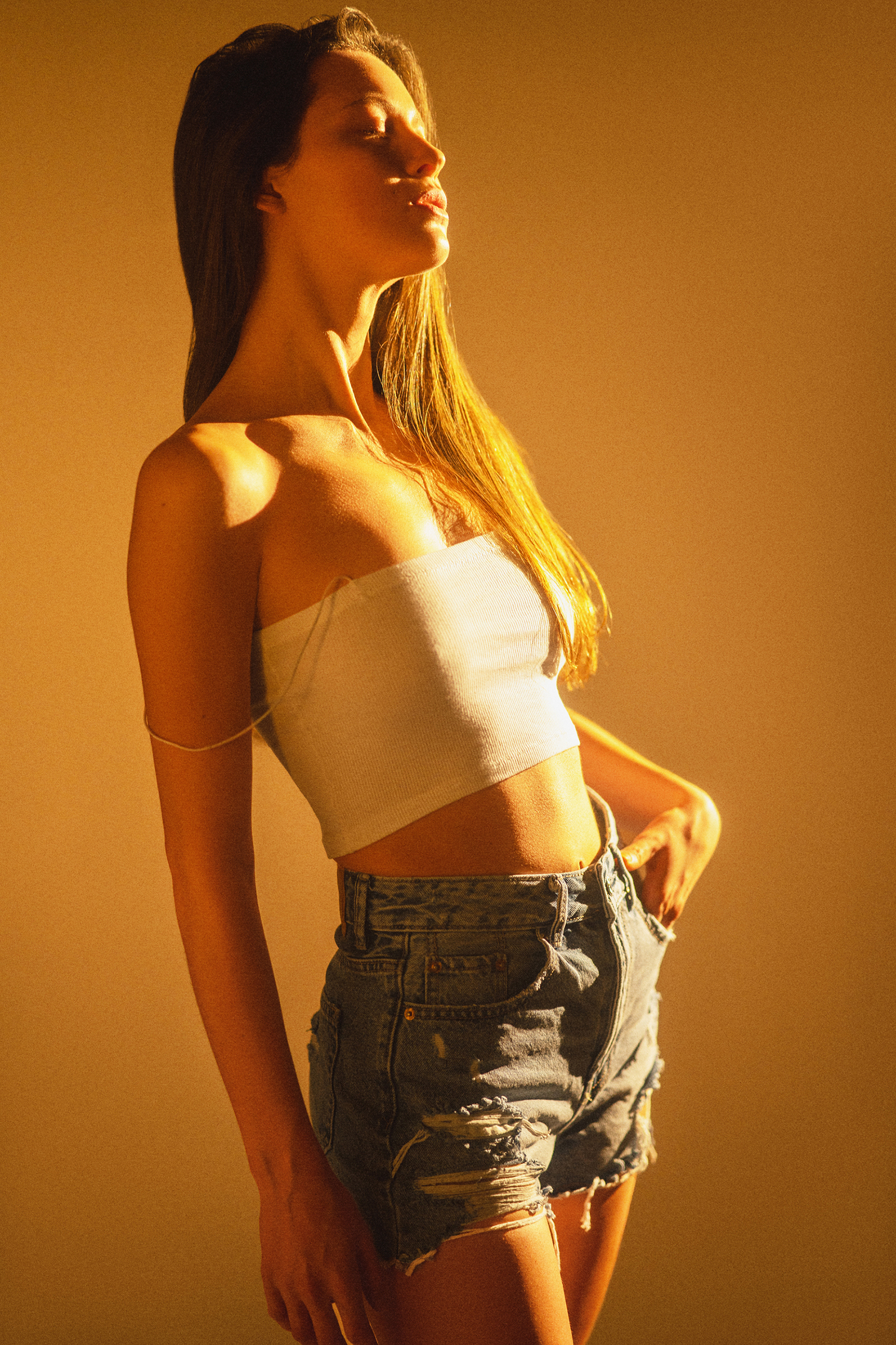 Editorial portrait of a woman in a white crop top and denim shorts posing in saturated amber light