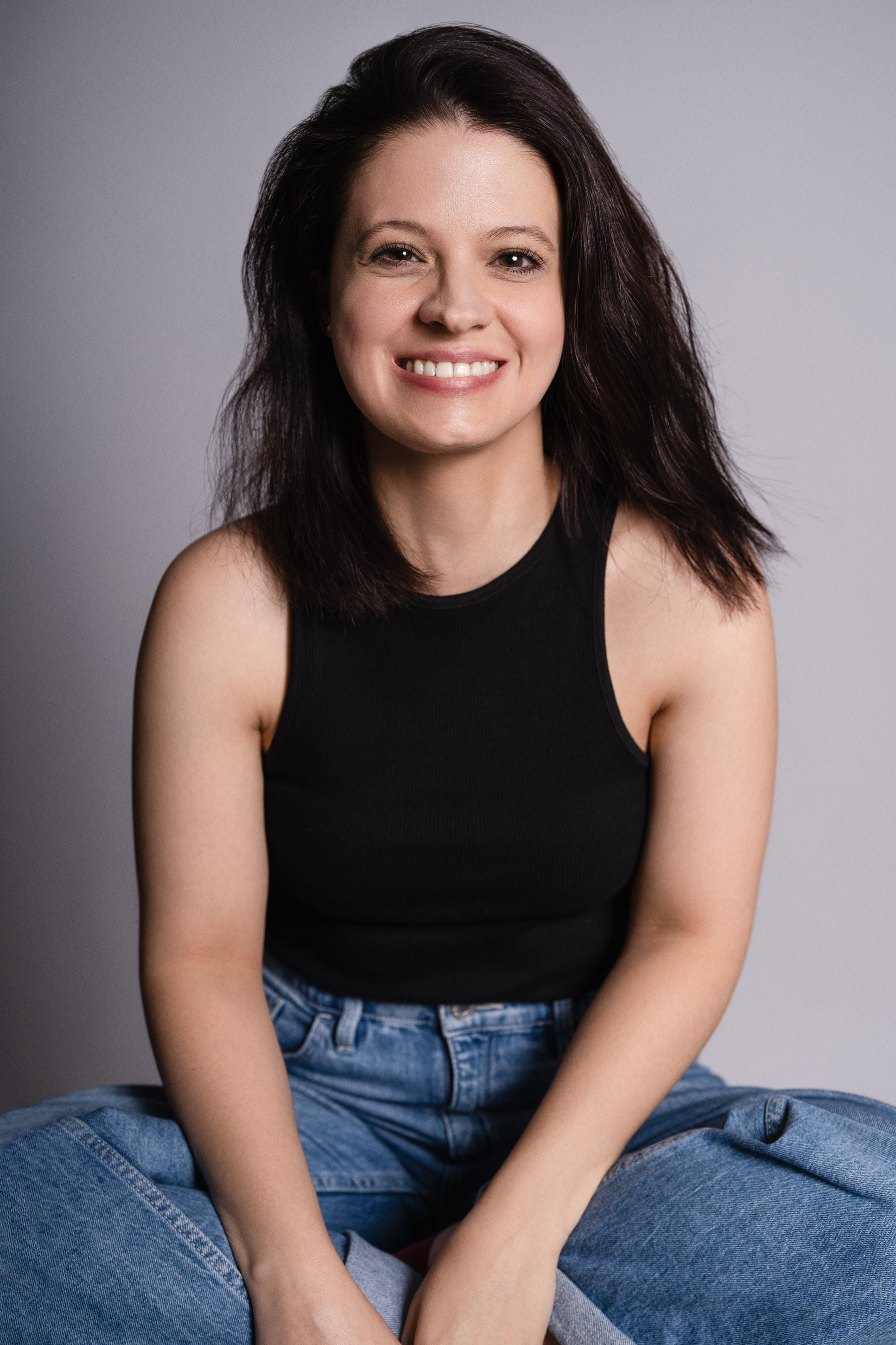 Relaxed studio portrait of a smiling woman in a black sleeveless top and blue jeans against a neutral gray background
