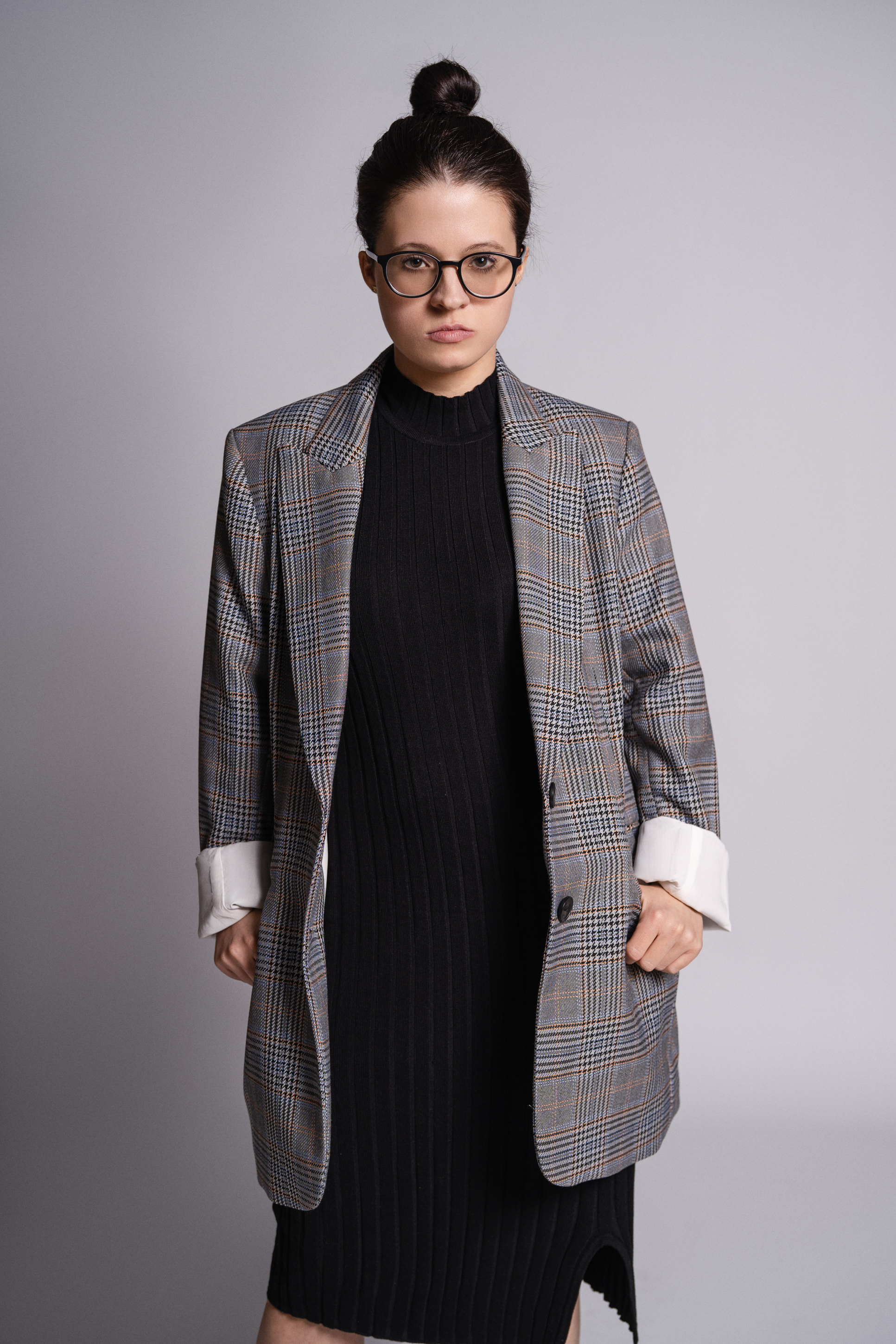 Full-length editorial portrait of a woman in an oversized checked blazer, black dress and glasses against a gray studio background