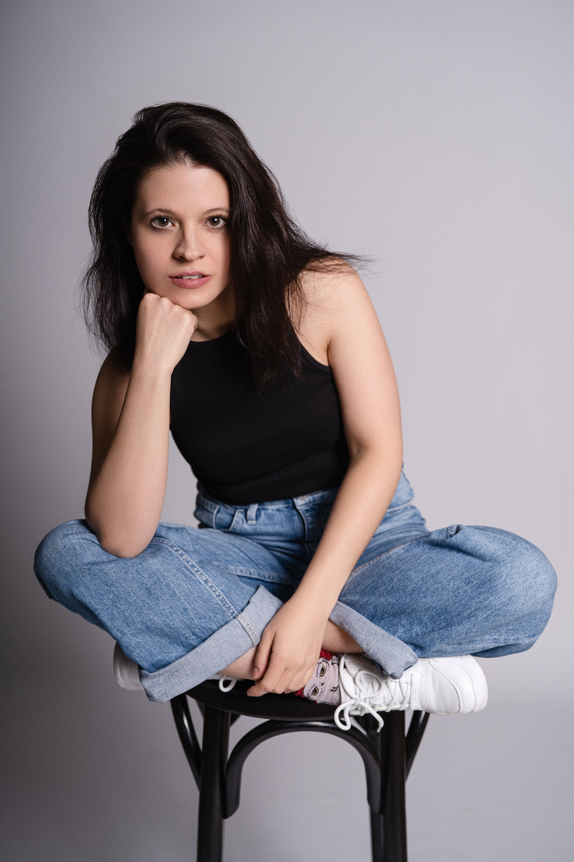 Casual studio portrait of a woman in a black top and loose blue jeans seated cross-legged on a chair