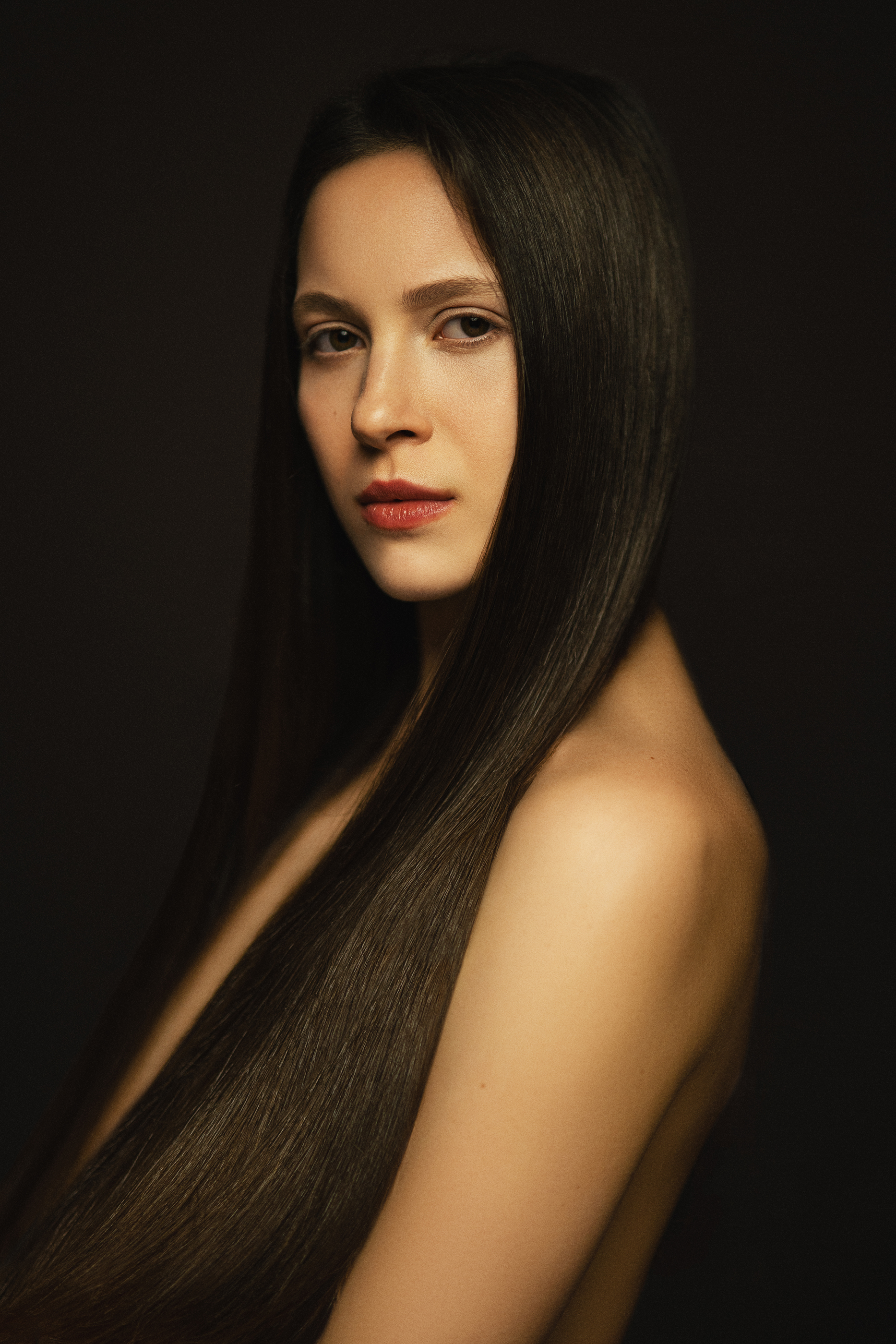 Close portrait of a woman with sleek long dark hair and a bare shoulder in soft low-key light