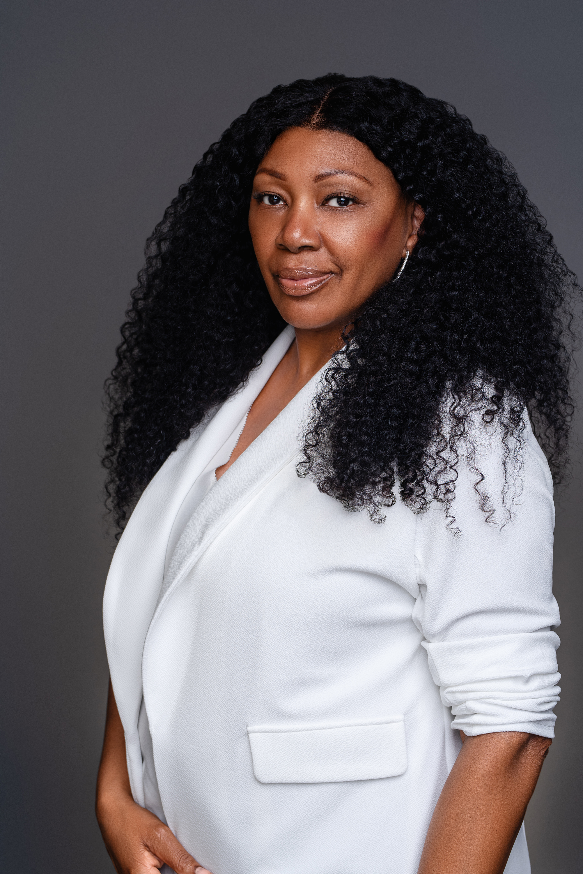 Studio personal branding portrait of a woman with long curly hair in a white blazer against a dark gray background