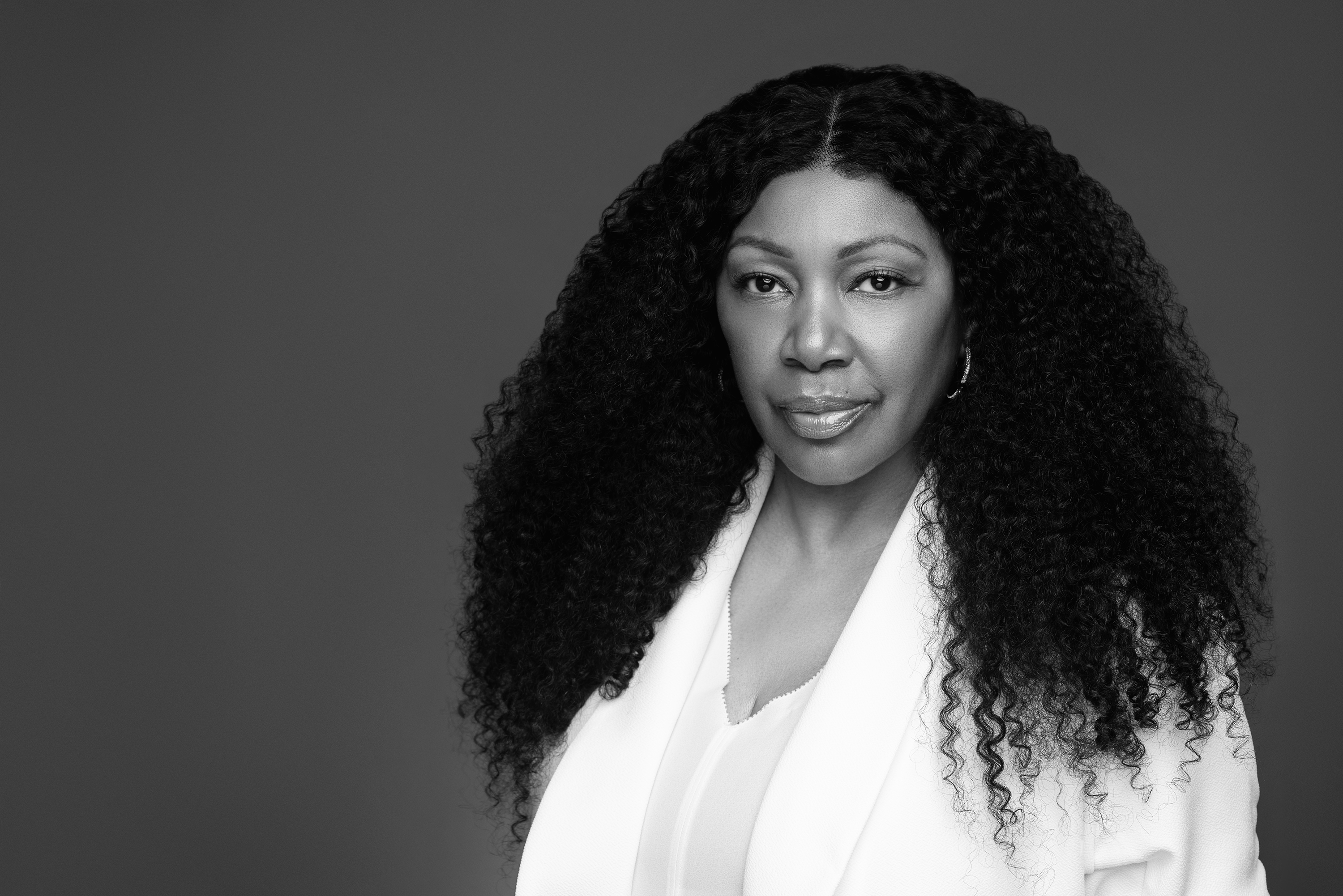 Black-and-white close portrait of a woman with long curly hair in a white blazer against a dark background