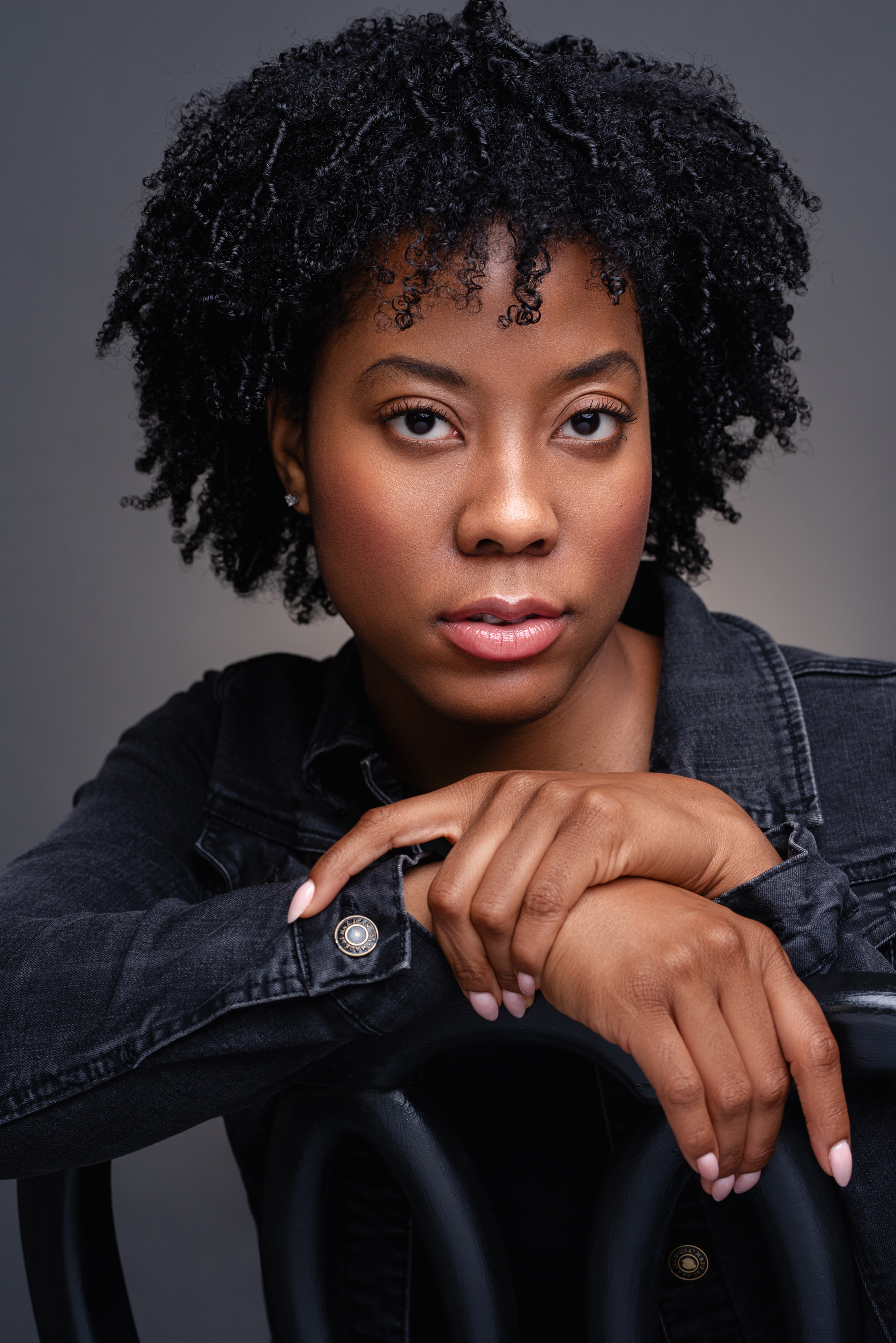 Studio headshot of a woman with natural curly hair in a dark denim jacket against a charcoal gray background