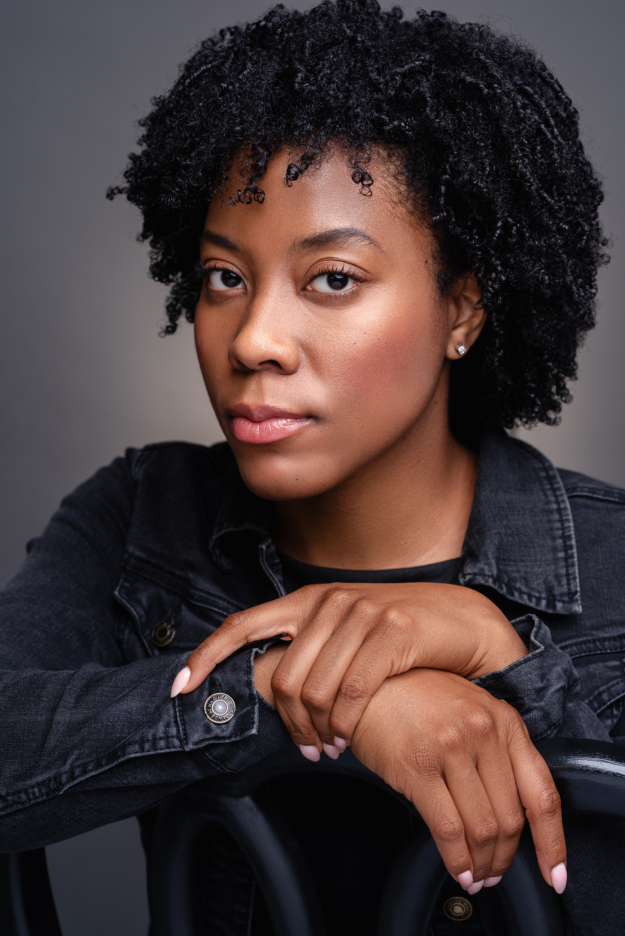 Close studio portrait of a woman with natural curly hair in a dark denim jacket against a charcoal background