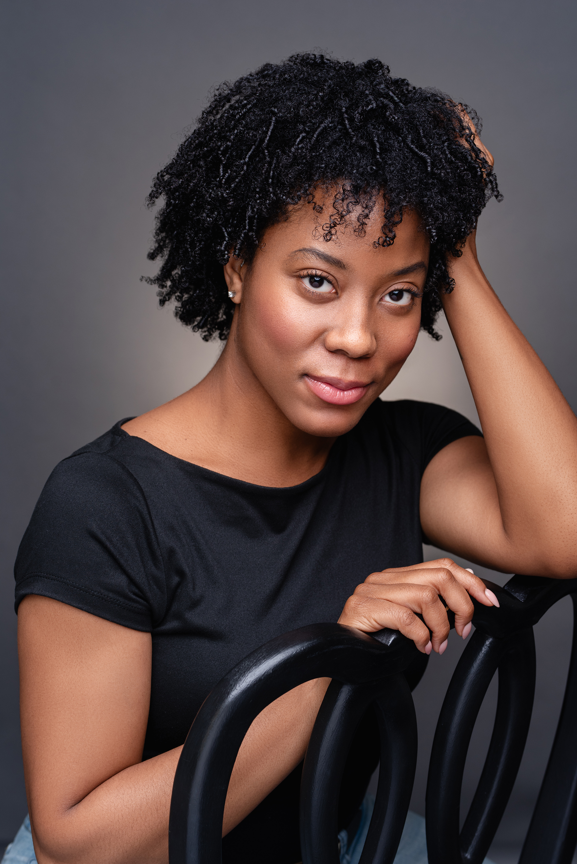 Studio portrait of a woman with natural curly hair in a black top seated against a soft gray background