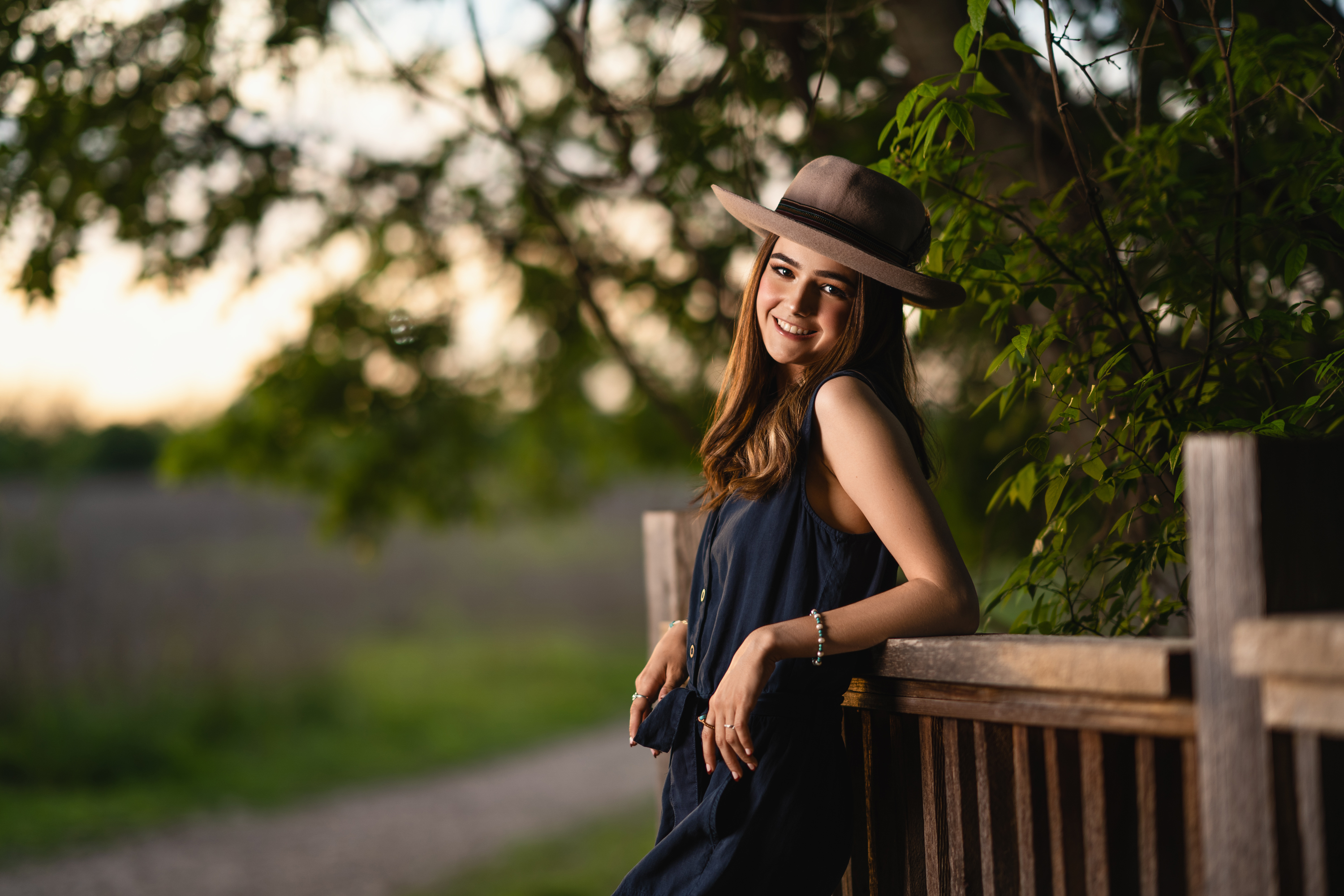 Outdoor senior portrait of a young woman in a dark outfit and wide-brim hat posing beside a wooden railing in warm evening light