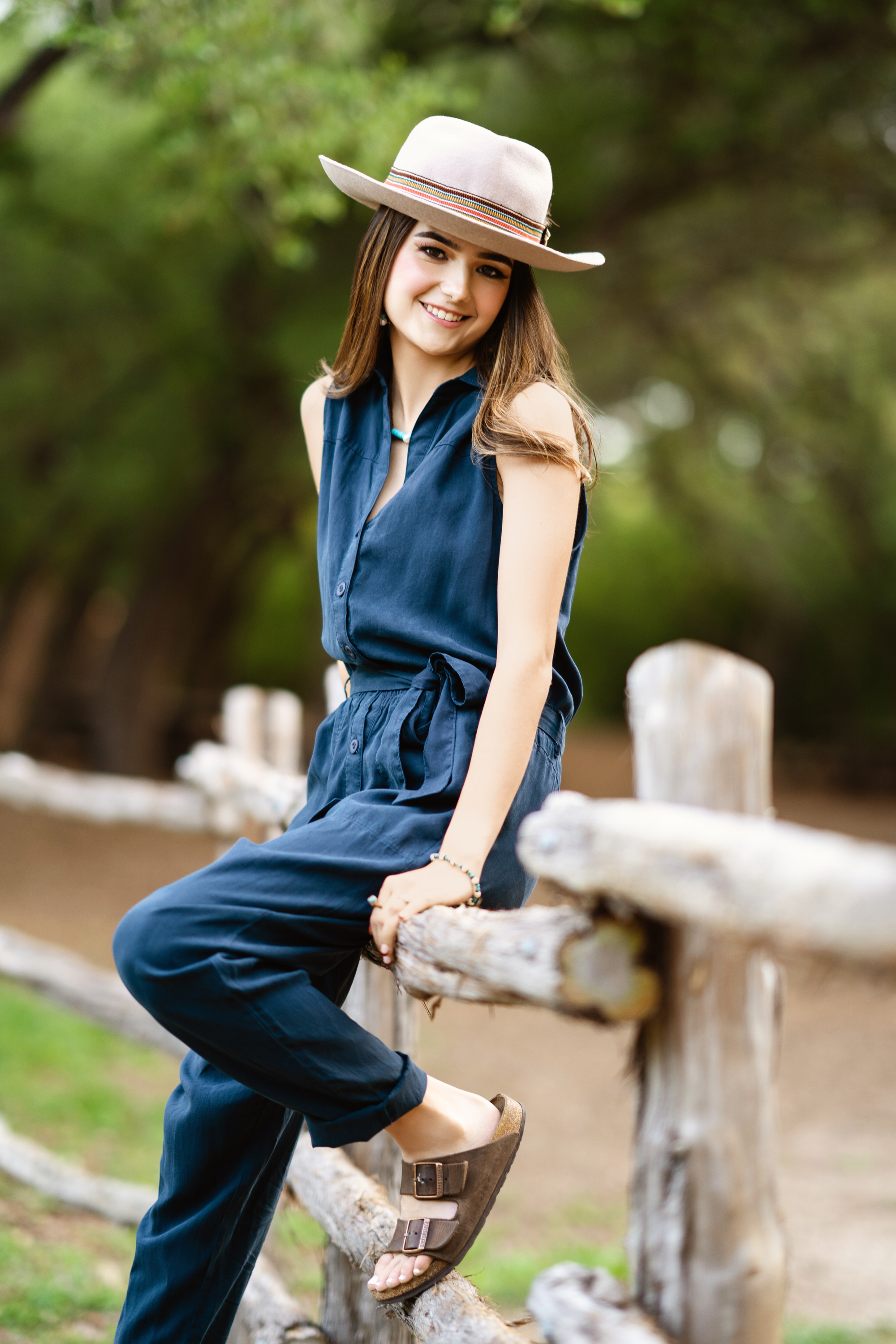 Outdoor senior portrait of a young woman in a blue jumpsuit and wide-brim hat seated beside a wooden railing