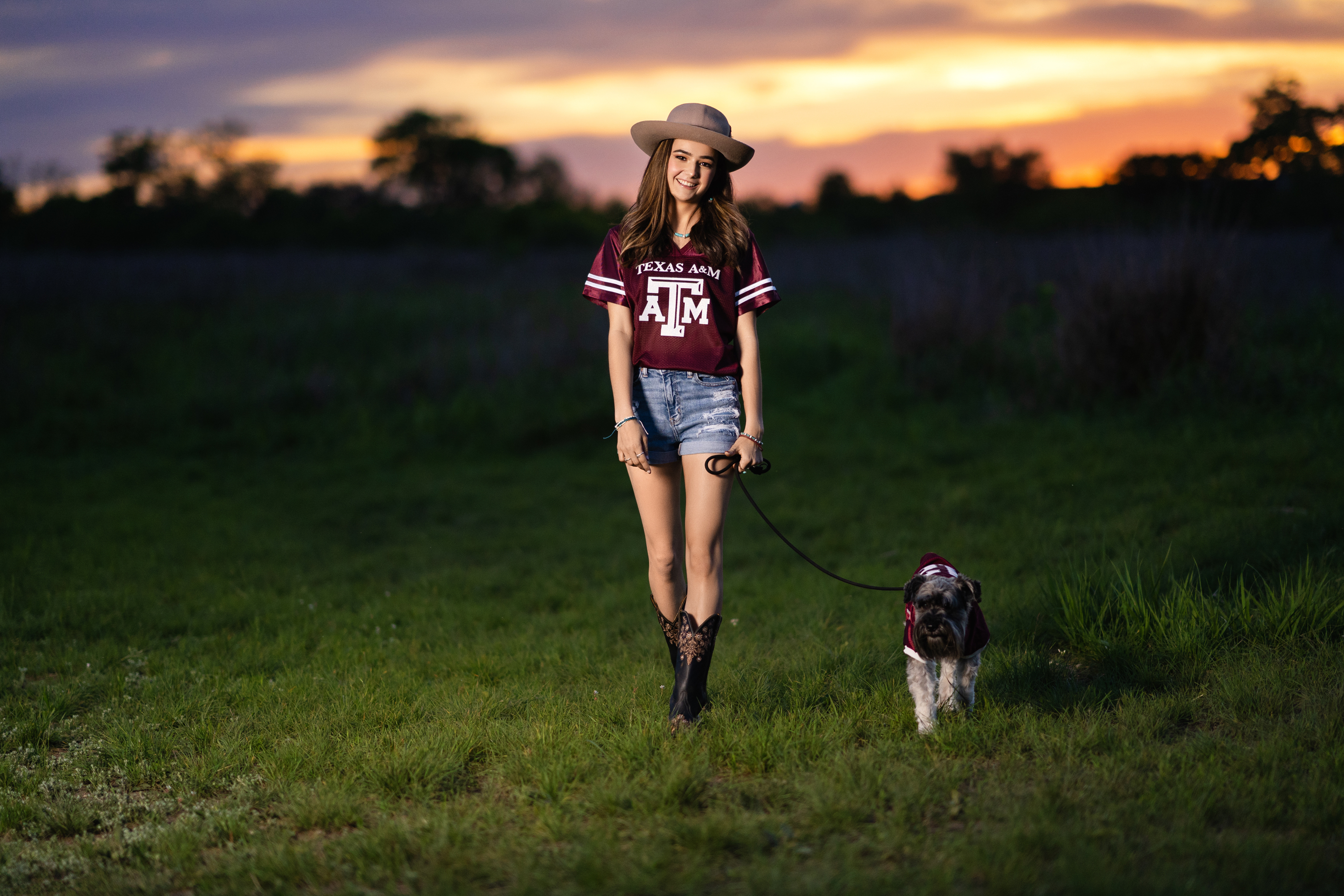Outdoor senior portrait of a young woman in a maroon college shirt walking through a field with her dog at sunset