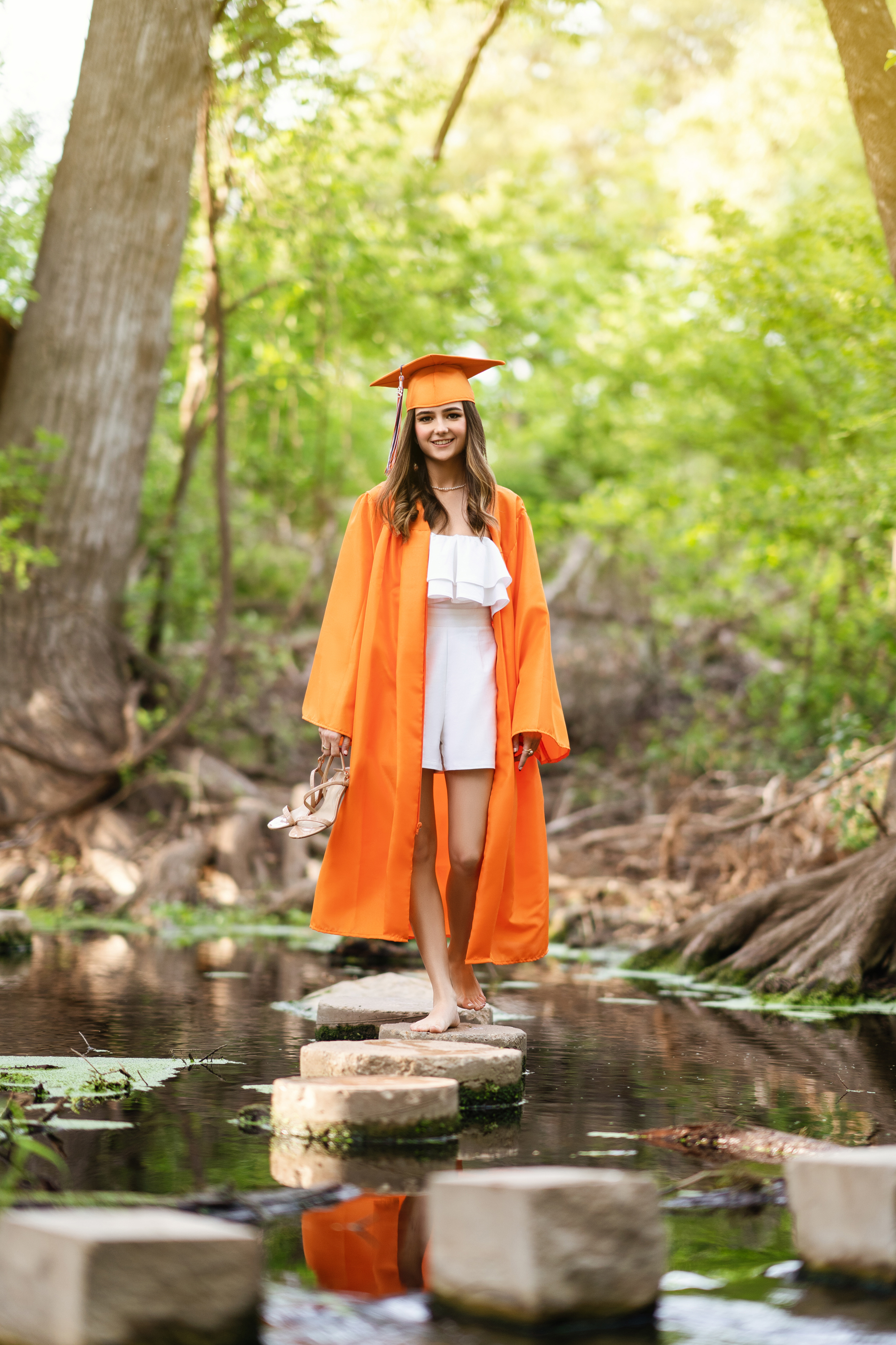 Graduation portrait of a young woman in an orange cap and gown standing beside a wooded creek